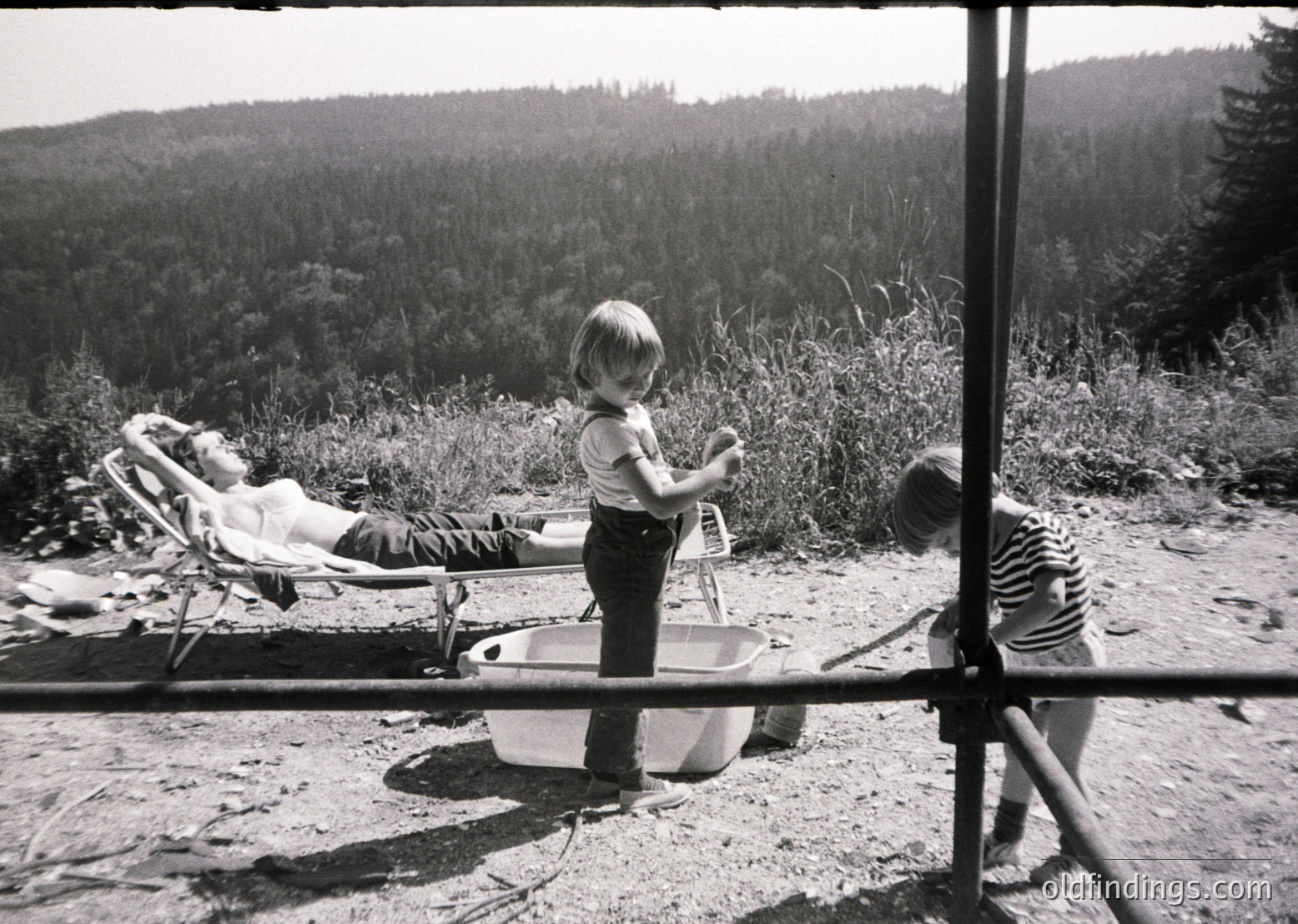 A mid-20th-century black-and-white snapshot captures a lakeside scene: a child in rolled-up pants stands beside a metal washbasin, while another reclines in a striped deck chair. Lush greenery and forested hills frame the background. Rustic wooden structures and a metal railing add to the vintage charm.