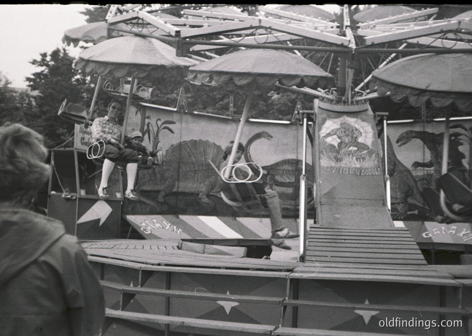 Mid-20th century carnival ride featuring a rotating dinosaur-themed structure with painted murals. Operators in vintage attire (1950s-60s) manage controls. Decorated with star motifs and bold lettering ("GATTA"). Crowd visible at base, suggesting public amusement park setting.