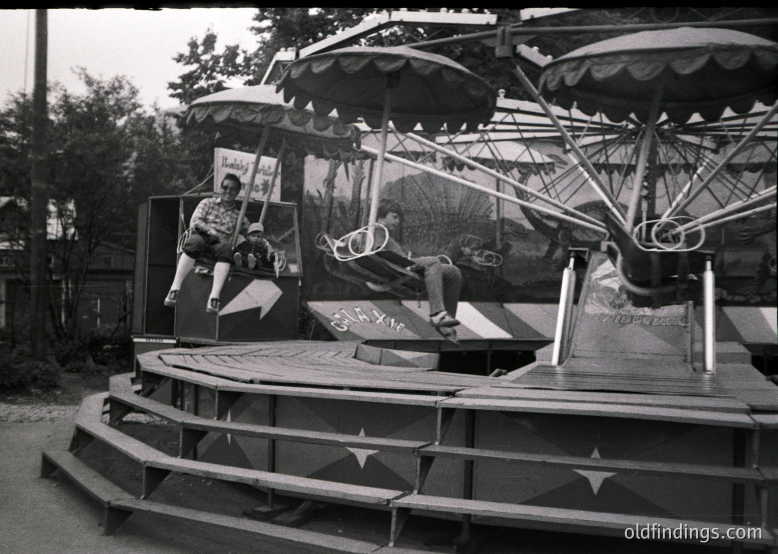 Vintage carousel with classic wooden seating and metal structure, featuring a "Tobacco" advertisement. Two seated riders pose mid-ride, one in shorts, one in a cap. Surrounding trees and fencing suggest a park setting. Likely mid-20th century (1950s-1960s).