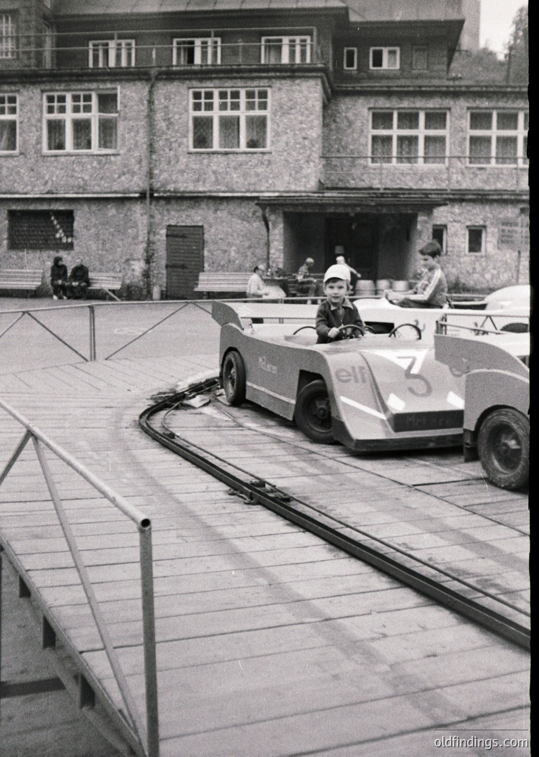 Vintage mid-20th century miniature train ride in a courtyard, featuring a child in a numbered "3" cart labeled "elf" on a track. Brick building with large rectangular windows in background.