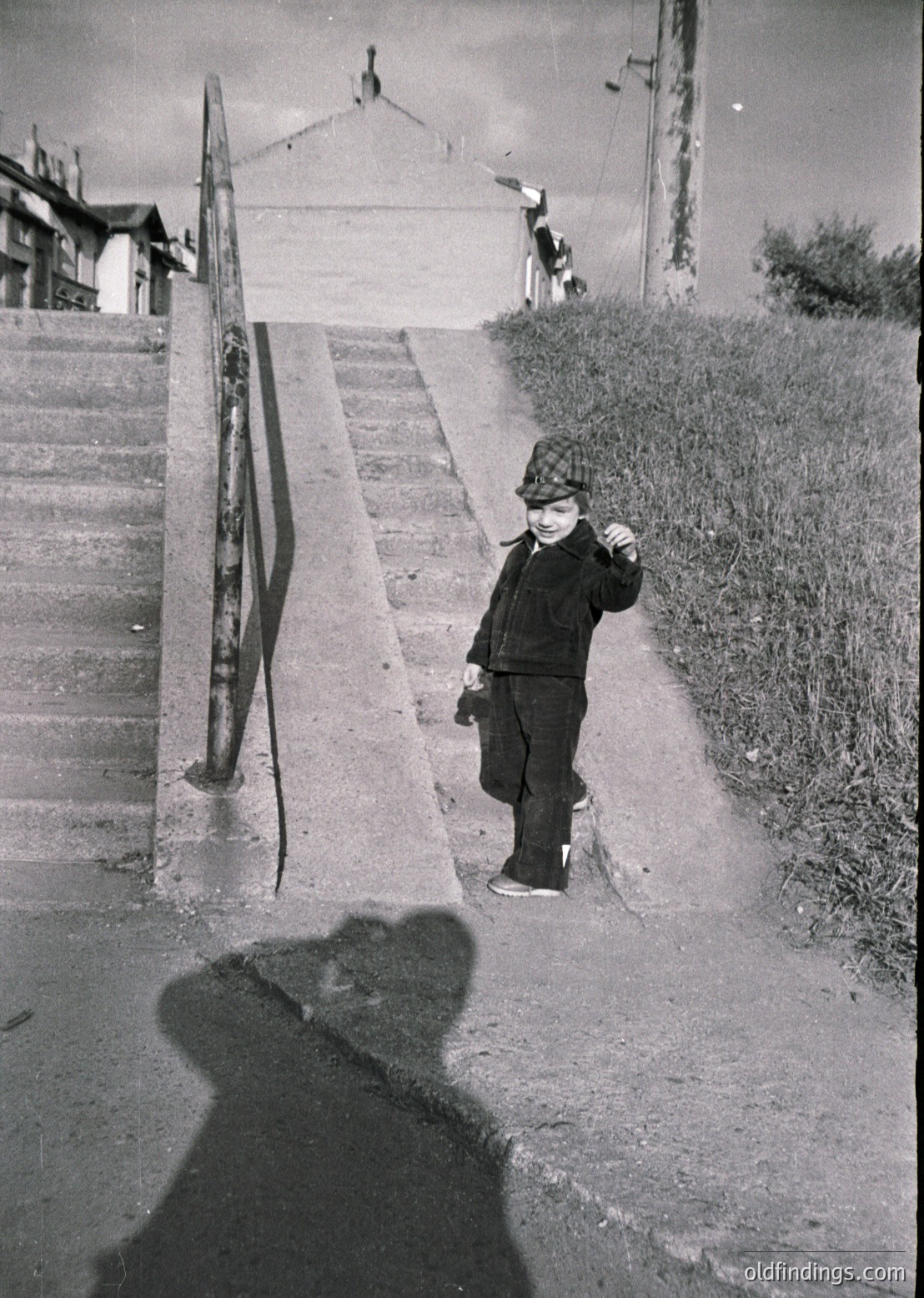 Mid-20th century street scene: Child in winter attire (hat, coat, boots) stands on concrete steps beside a rusted metal railing. Shadow cast by overcast sky. Background shows weathered buildings and utility poles. Urban residential setting, likely post-WWII era.