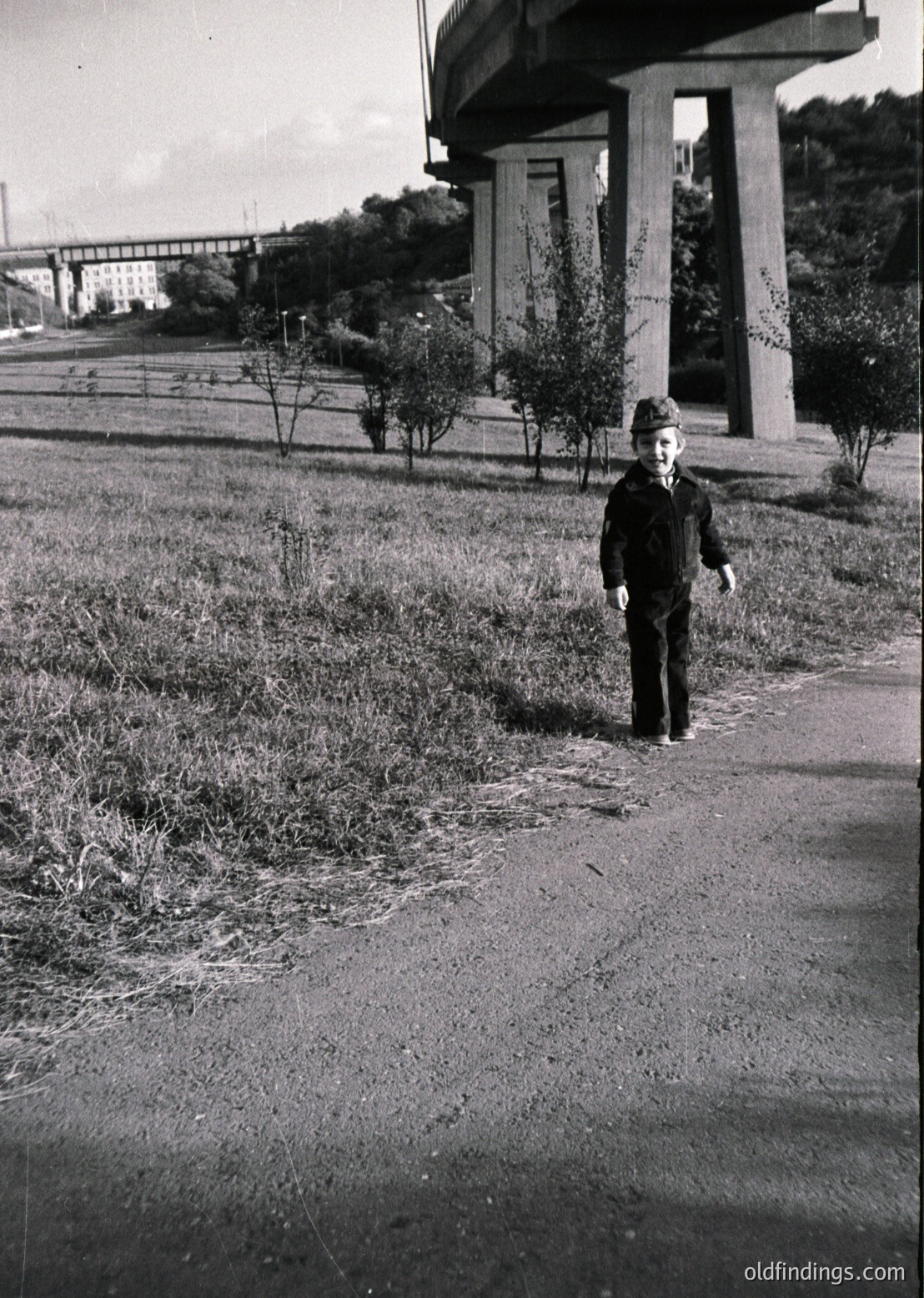 A young boy in mid-20th century attire stands on a paved path beside a concrete overpass, surrounded by overgrown vegetation. His cap and knee-length pants suggest or urban/suburban setting. Industrial infrastructure and greenery hint at post-war reconstruction or early development.