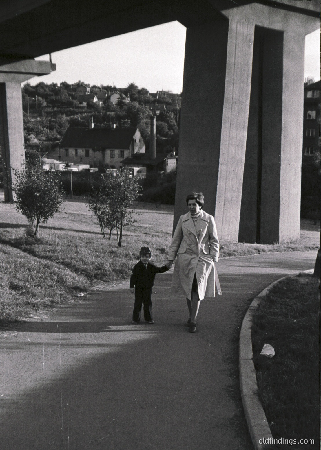 A woman in 1950s-era coat and hat leads a young boy under a modernist concrete overpass, flanked by small trees. Mid-century urban landscape with residential buildings in background.