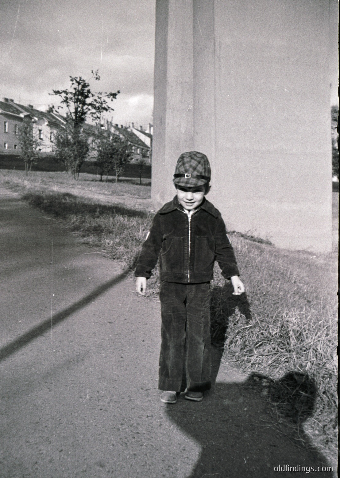 Mid-20th century street scene: Child in 1950s-style jacket and checkered cap holding a small bundle, walking beside a concrete wall. Residential buildings and bare trees in background suggest urban setting. Classic candid street photography.