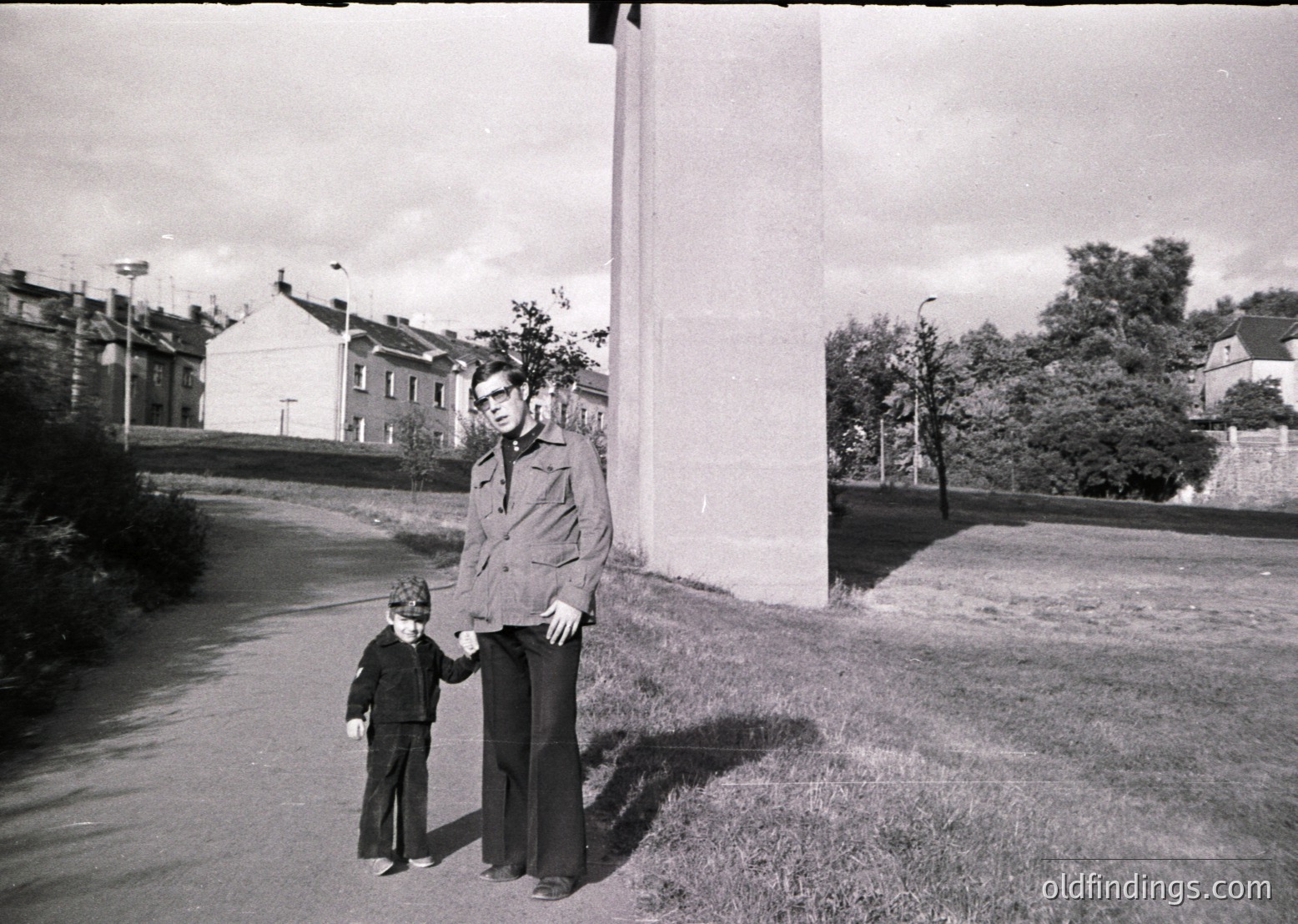 Mid-20th century street portrait: man in 1950s-style jacket and glasses holds child in formal attire beside a tall concrete pillar. Residential buildings and trees in background suggest urban/suburban setting.