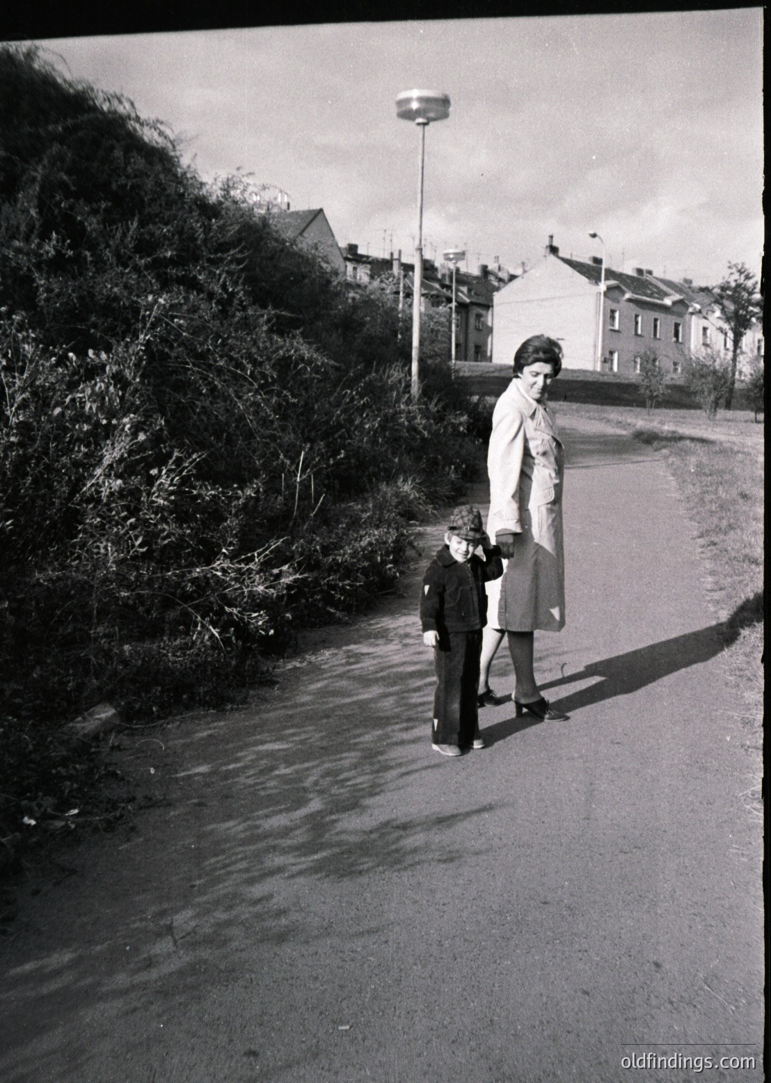A woman in a long coat and a child in a sweater and shorts walk along a wet, paved road beside dense shrubbery. Mid-20th century residential buildings and a streetlamp frame the scene.
