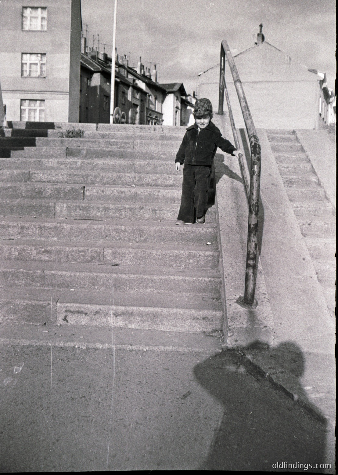 A child in early 20th-century attire descends a wide, concrete staircase flanked by metal railings. Surrounding buildings feature Soviet-era architecture with flat roofs and simple facades. The scene suggests an urban setting, likely Eastern Europe, during the mid-1900s. [Child descending Soviet-era staircase in urban setting, 1950s–1960s ]