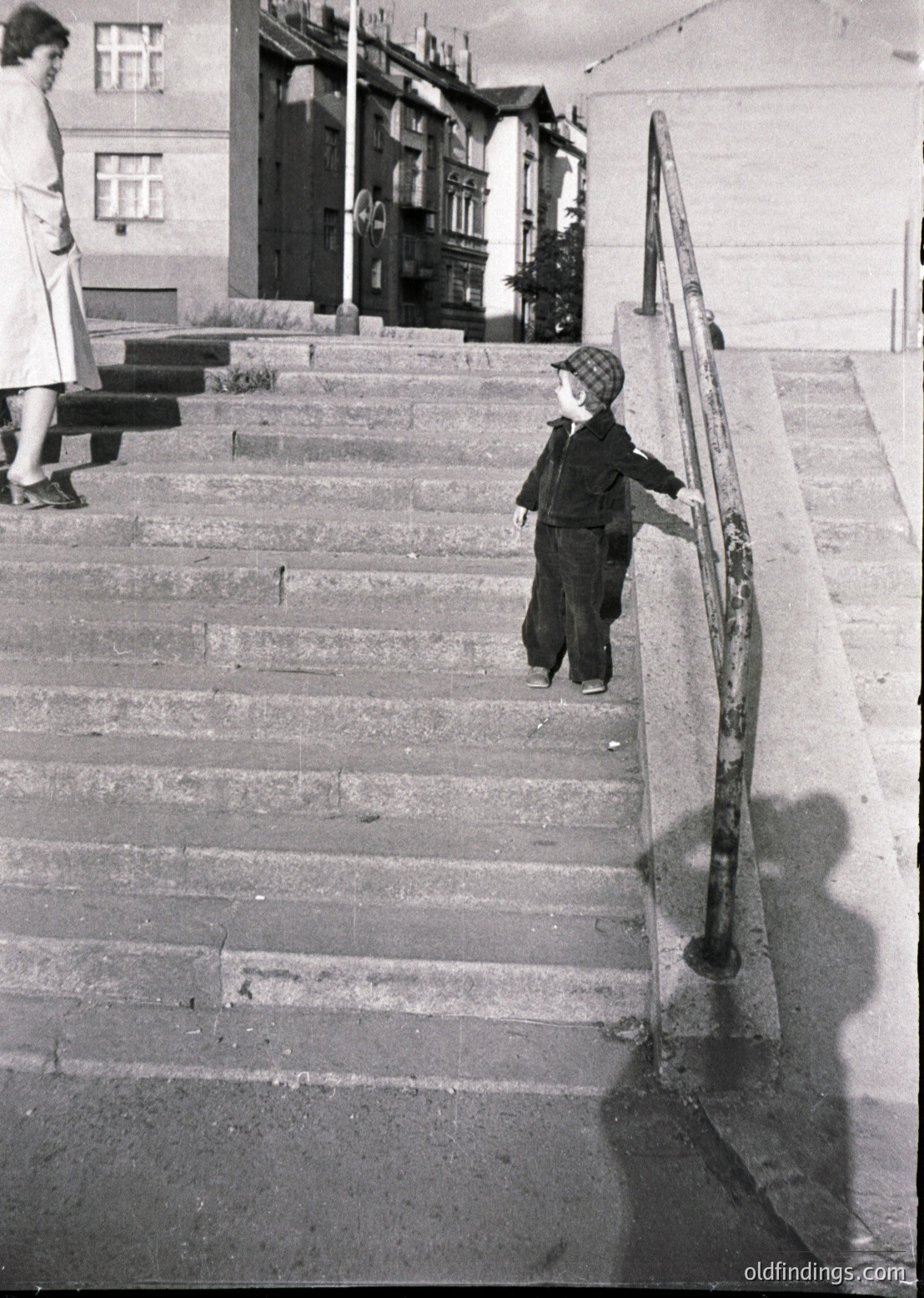Mid-20th century black-and-white street scene: Boy in sweater and cap descends concrete stairs with metal railing, holding handrail. Adult in long skirt stands on upper landing. Multi-story residential building with balconies and potted plants in background. Urban setting, likely Eastern European.