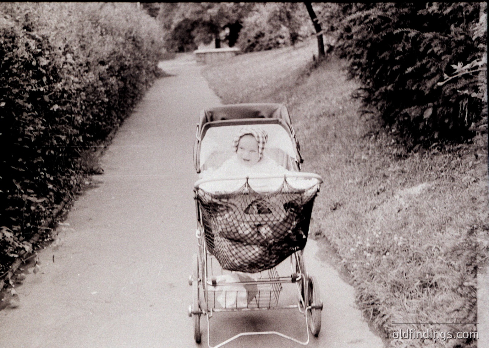 Vintage black-and-white photo of a child in a classic wicker pram on a tree-lined pathway, mid-20th century. Basket details suggest 1950s-1960s design.