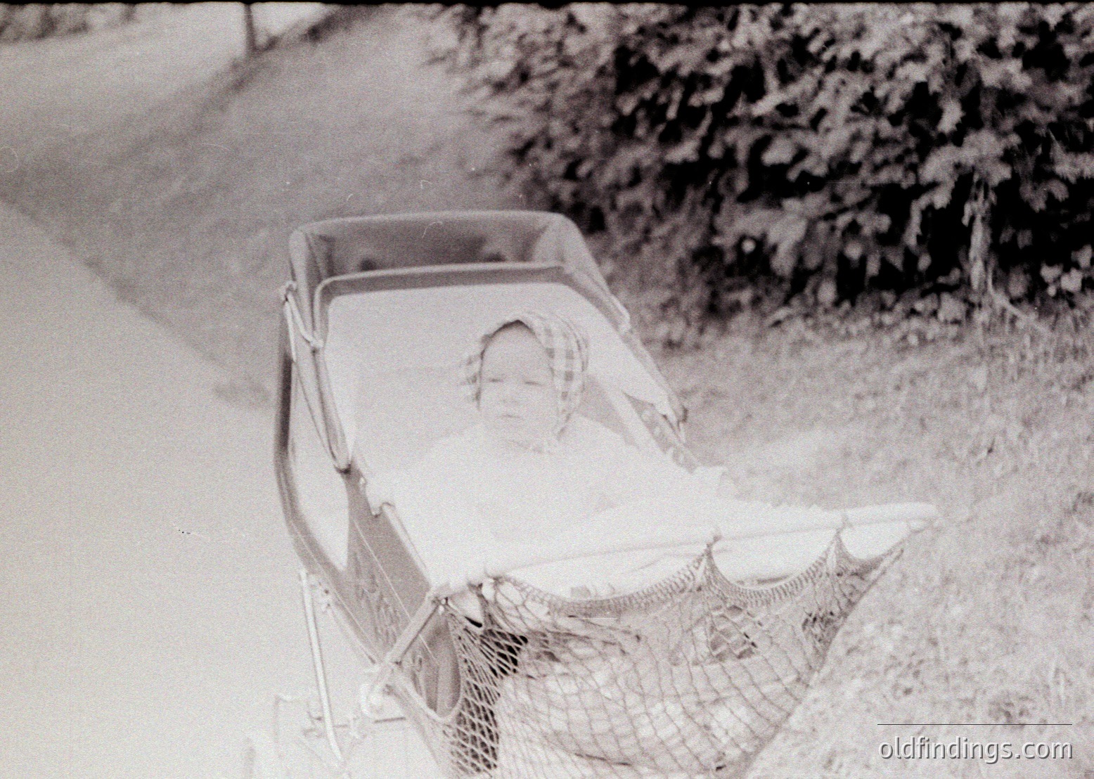 Vintage black-and-white photo of a child reclining in a vintage pram with a mesh canopy, likely mid-20th century. The setting appears to be a rural or suburban pathway surrounded by greenery. The child’s relaxed posture suggests a moment of rest or leisure.