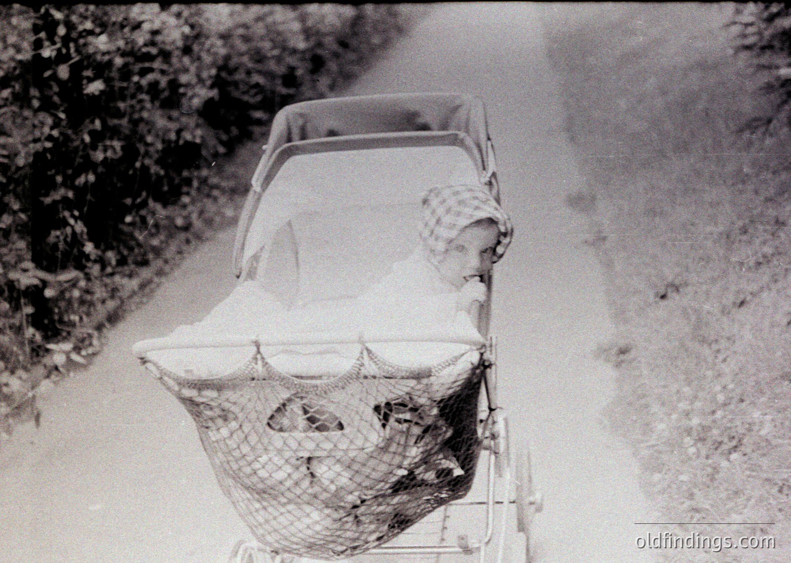Vintage black-and-white photo of a baby in a mesh-woven pram, pushed along a rural dirt road. The infant wears a patterned bonnet and sleeps under a light blanket. Mid-20th century design reflects practical, utilitarian parenting styles.