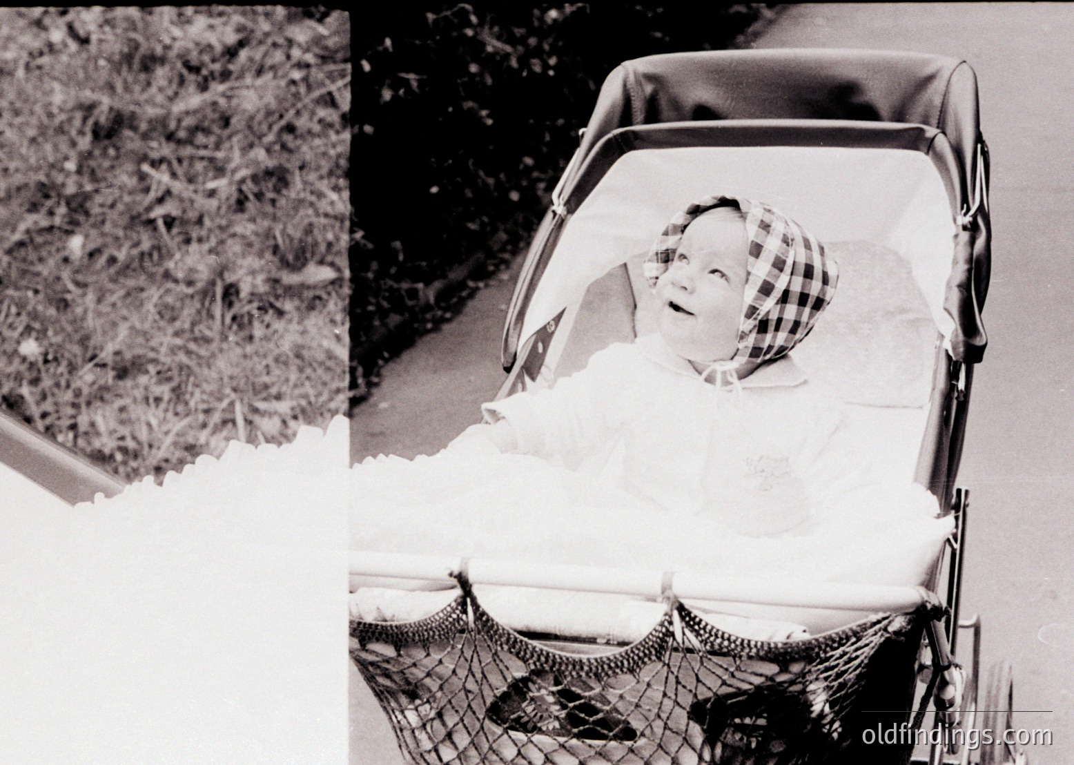 Vintage black-and-white photo of a child in a vintage pram with a woven mesh basket, likely mid-20th century. The child wears a checkered bonnet and a long-sleeve dress, seated outdoors near foliage. Classic mid-century design and nostalgic family imagery.