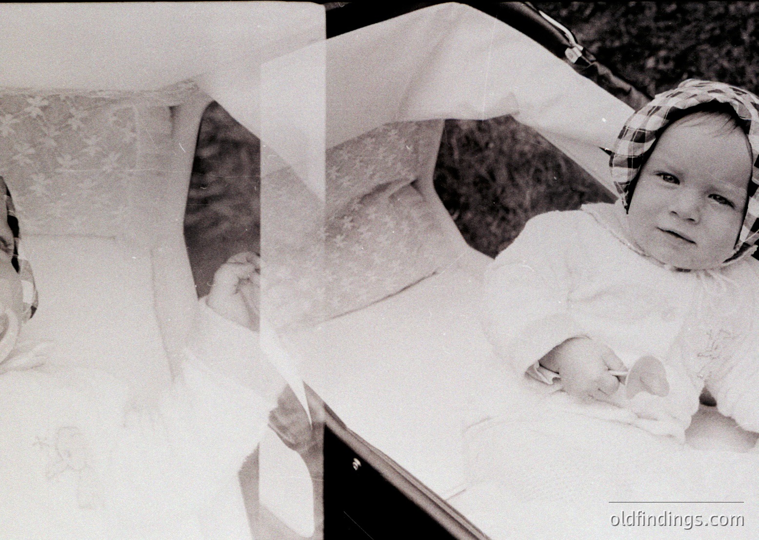 Vintage black-and-white photo of a baby in a white sleeper, lying on a flat surface with blurred background. The infant’s head is slightly tilted, and a headband or hat is visible. Likely mid-20th century family portrait.