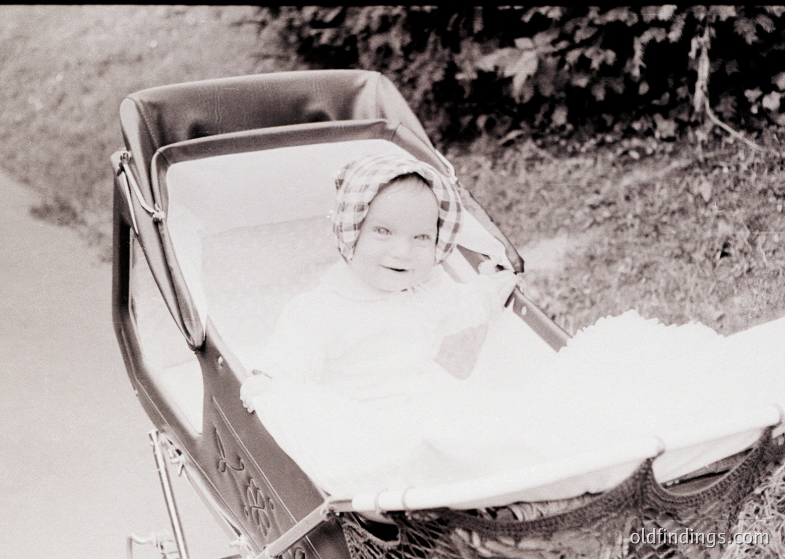 Vintage black-and-white photo of a child in a vintage metal-framed stroller, outdoors on a dirt path. The child wears a checkered headscarf and light clothing, suggesting mid-20th century. Surrounding foliage hints at a rural or park setting.