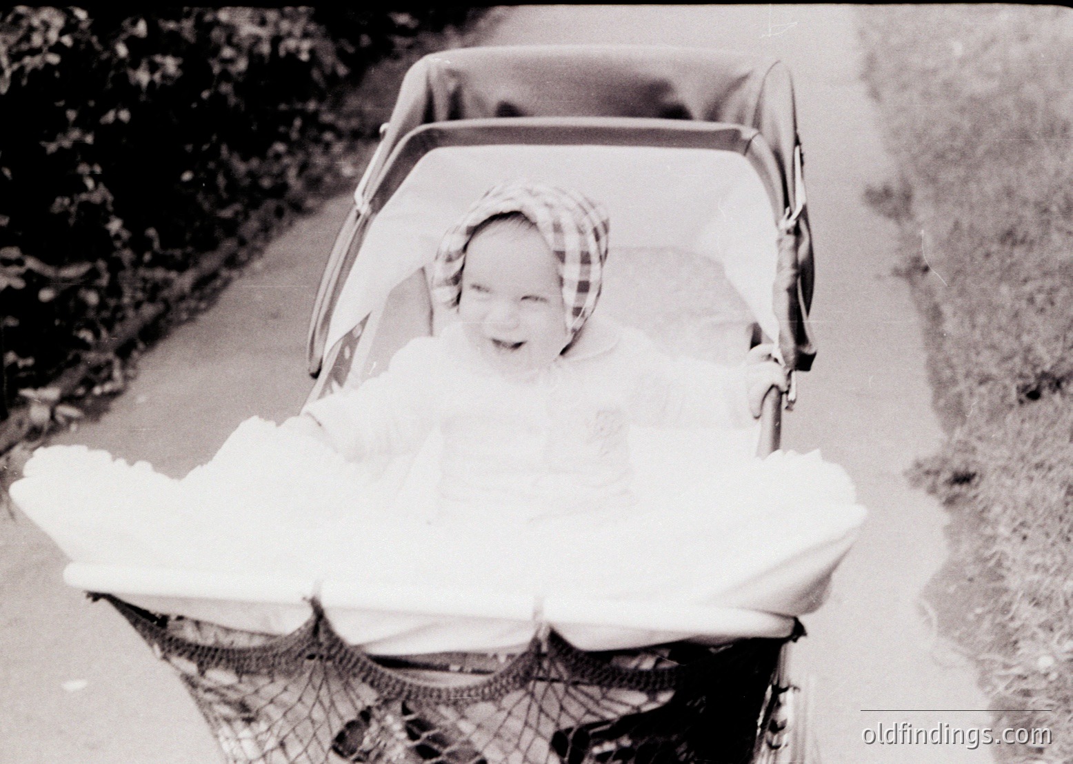Vintage black-and-white photo of a child wrapped in a thick blanket, seated in a classic wicker pram with a mesh front. The child wears a checkered headscarf and smiles directly at the camera. Likely mid-20th century, urban or suburban setting.