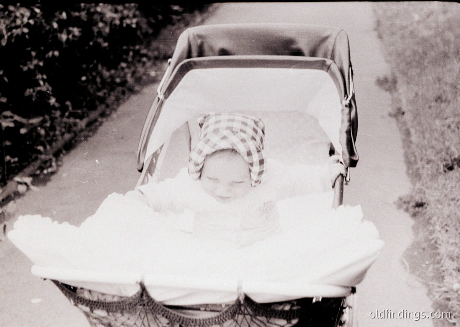 Vintage black-and-white photo of a child in a classic stroller, wrapped in a blanket, with a checkered headscarf. Urban pathway with greenery on one side, suggesting mid-20th century daily life.