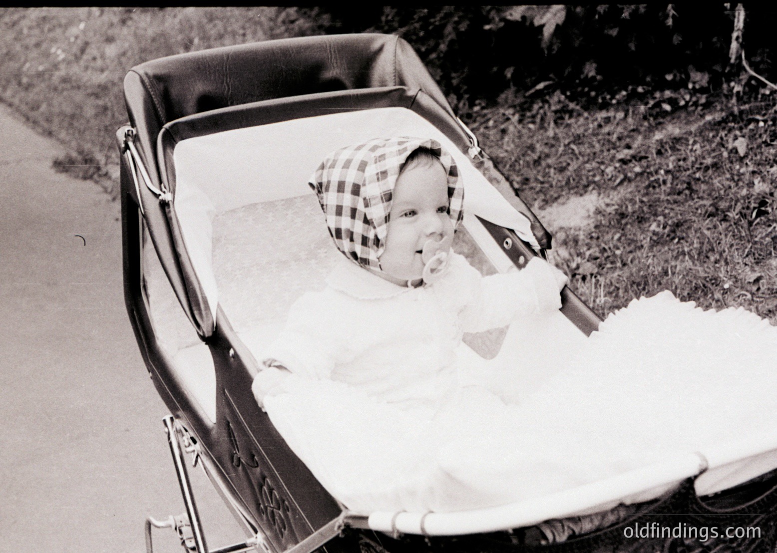 Vintage black-and-white photo of an infant in a classic metal-framed pram, wearing a checkered cap. Soft blanket and padded seat suggest mid-20th century design. Grass and pavement hint at an outdoor setting, likely residential.