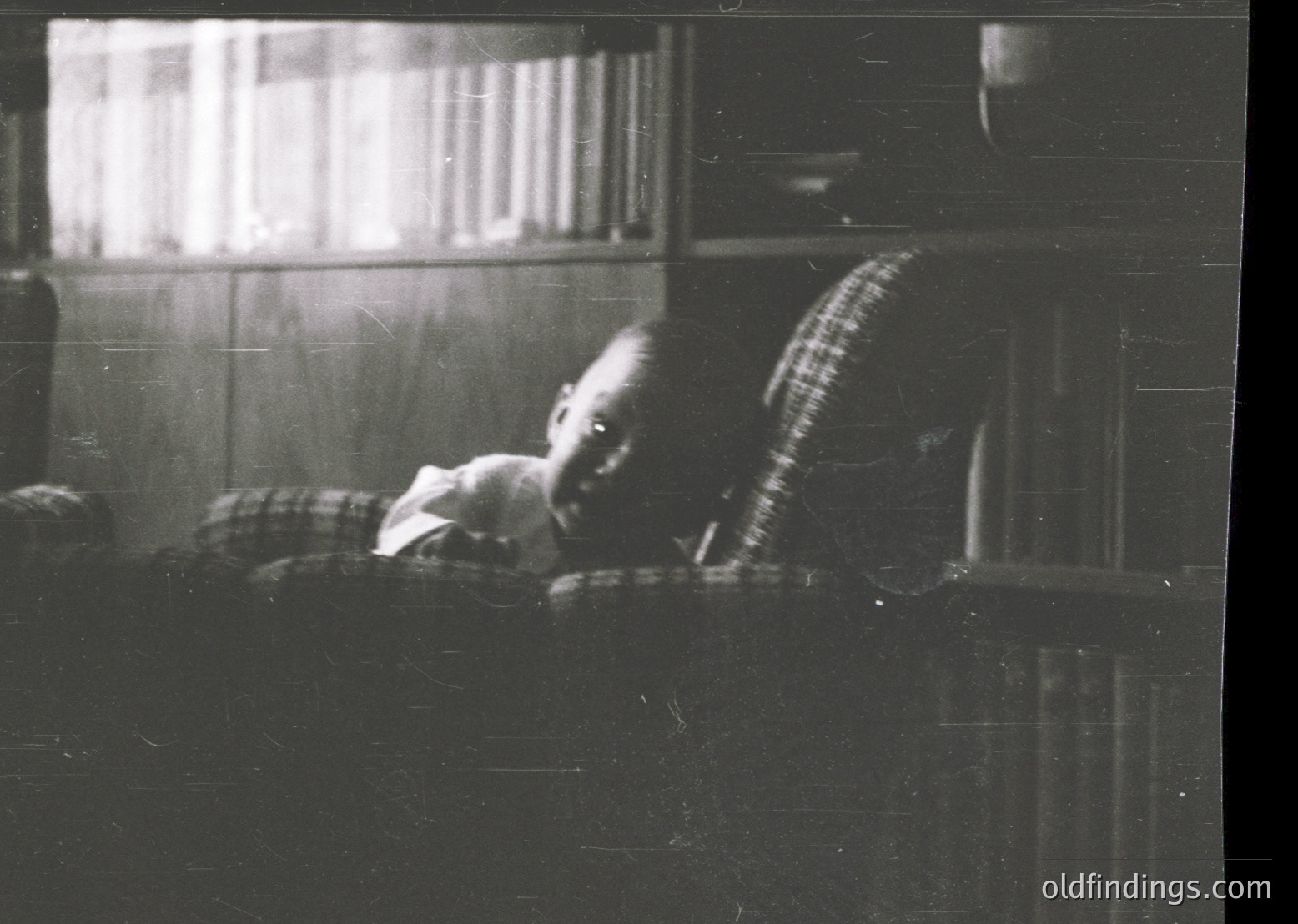 Vintage black-and-white photo of a child resting on a window sill, draped in a striped blanket. Indoor setting with visible brickwork and a partial view of a bed. Likely mid-20th century domestic interior.