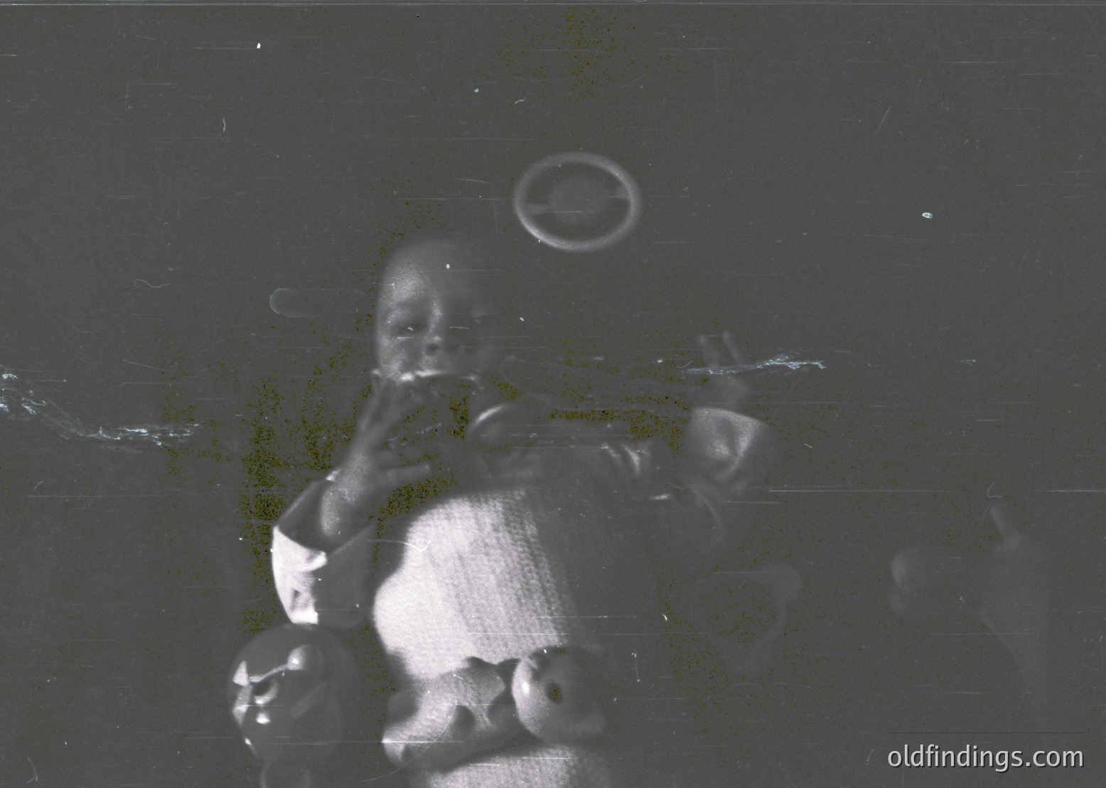 Vintage black-and-white photo of a young child in a seated position, holding a small object to their mouth. The grainy texture and lighting suggest early 20th-century photography. The child appears to be indoors, possibly in a dimly lit room.