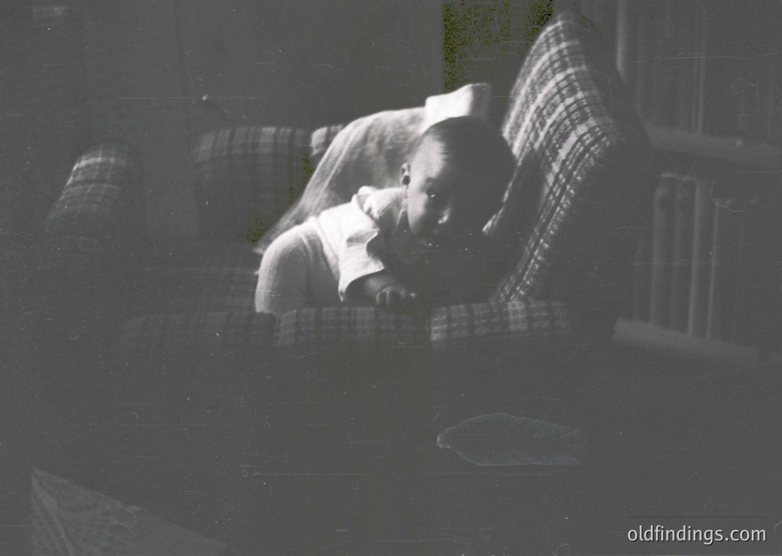 Vintage black-and-white photo of a child sleeping on a plaid blanket, likely mid-20th century. Soft focus and warm lighting suggest a cozy indoor setting, possibly a bedroom. The blurred background hints at a window or door with light filtering through.
