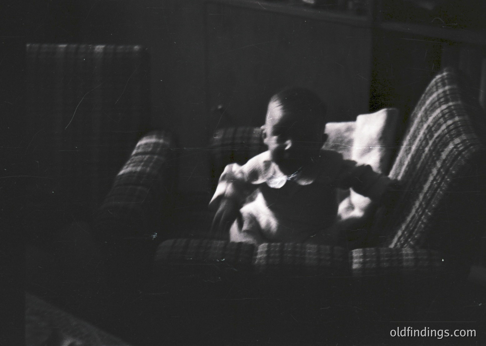 Vintage black-and-white photo of two figures seated on a striped armchair, blurred motion suggests movement or candid capture. Indoor setting with partial view of a curtained window. Likely mid-20th century domestic scene.