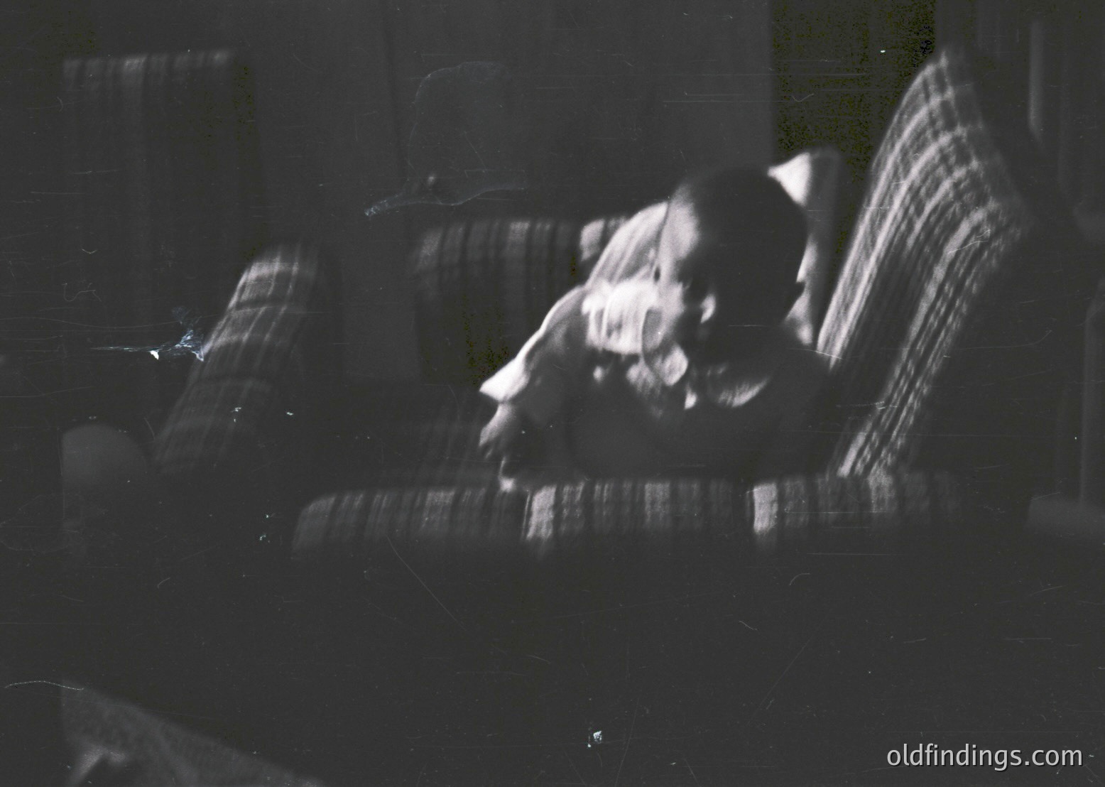 Vintage black-and-white photo of a person reclining on a striped armchair, holding a cigarette. Soft focus and grainy texture suggest mid-20th century style, likely 1950s–1960s. Indoor setting with partial view of a window and curtain.