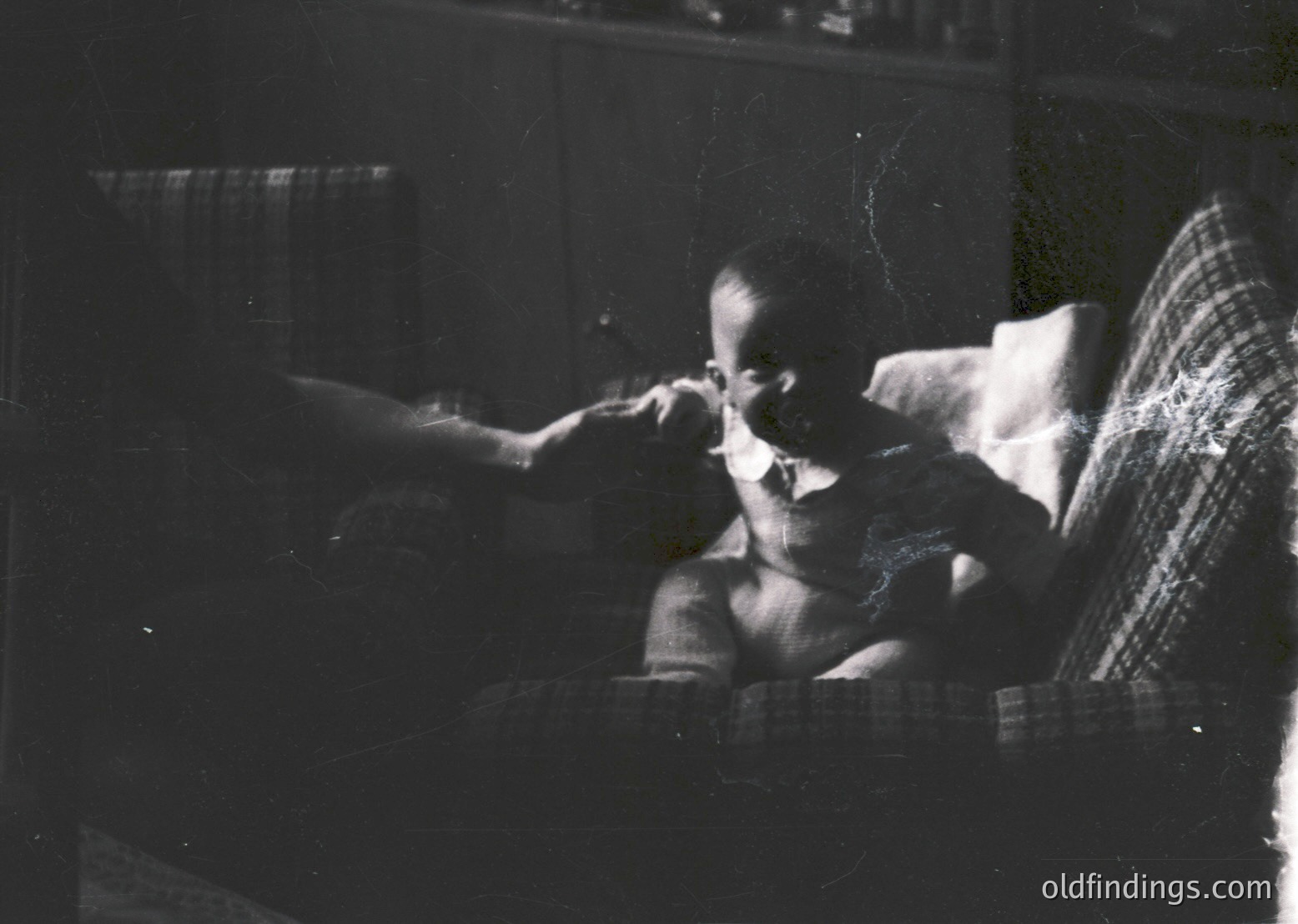 Vintage black-and-white portrait of a seated child brushing teeth in a dimly lit indoor setting, likely mid-20th century. Checkered cushion and bookshelf in background suggest a domestic, mid-class home.