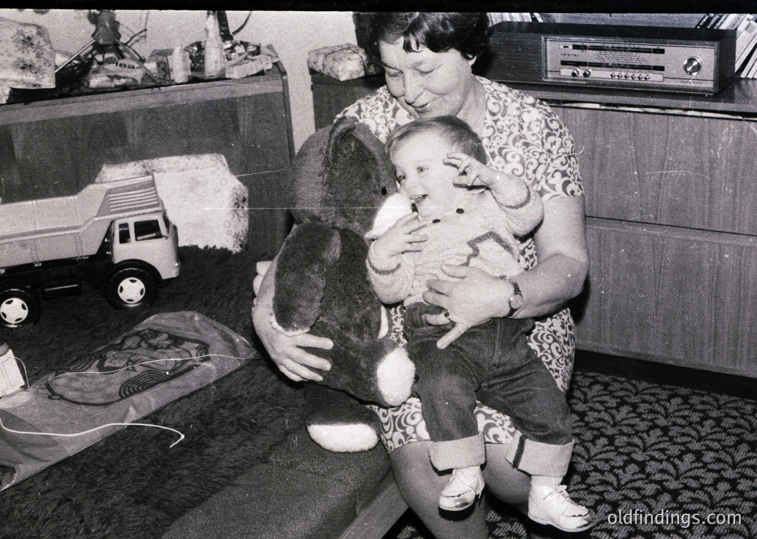 A woman cradles an infant while holding a stuffed animal, likely mid-20th century (1960s–1970s). Surrounding items include a toy truck, vintage radio, and patterned flooring. Warm, nostalgic family moment.