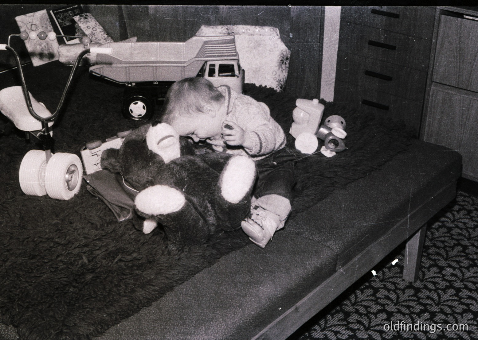 A child in a 1960s-70s hospital gown plays with a toy on a metal bed, surrounded by medical equipment like a stethoscope and IV pole. The vintage black-and-white photo captures a candid moment of childhood resilience amid medical care.