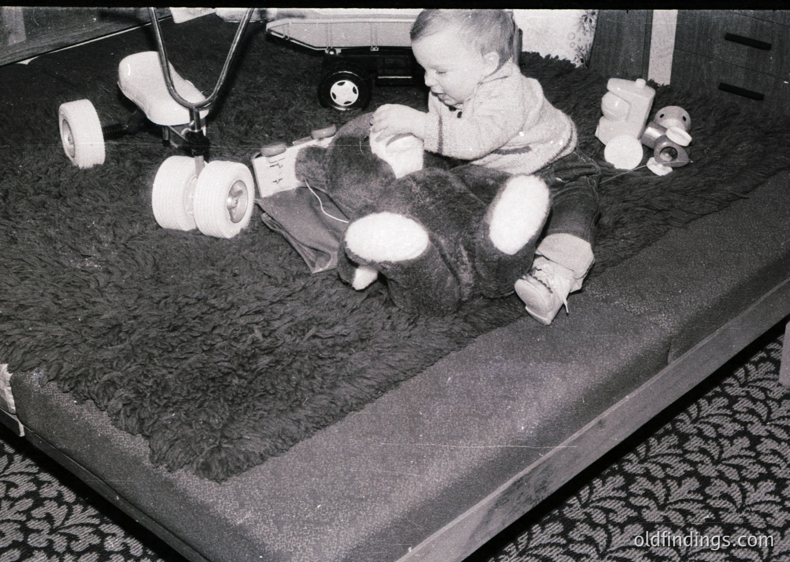 A toddler in mid-1970s medical leg braces plays on a padded hospital bed, surrounded by rolled bandages and a stuffed cow toy. The setting suggests a pediatric rehabilitation unit.
