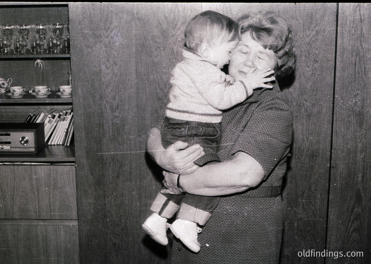 A tender mid-20th-century moment: an adult cradles a baby in a cozy indoor setting. The woman’s sweater and the child’s striped shirt suggest 1950s–1960s fashion. Shelves behind display vintage teacups, books, and a radio, hinting at mid-century domestic life.