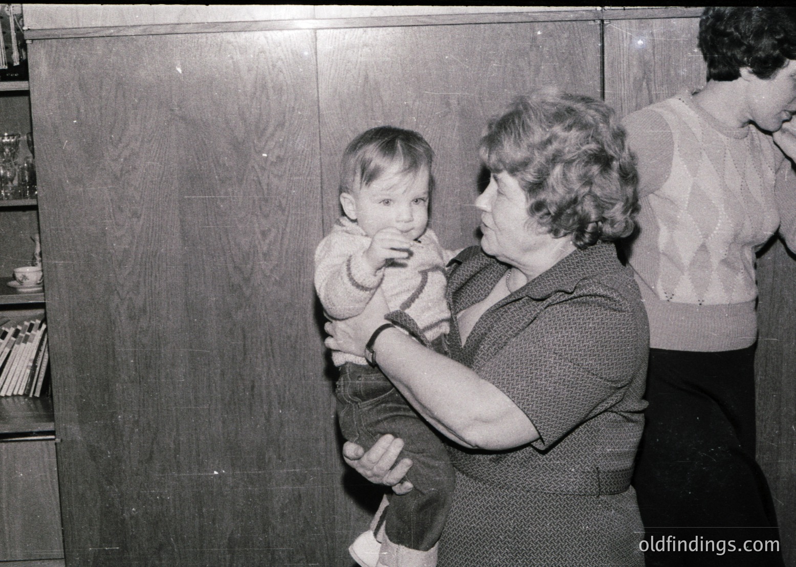 A woman cradles a toddler in a 1960s-70s indoor setting, likely a school or community hall. Wooden cabinets and shelves with books or supplies in the background suggest an institutional environment. The woman’s patterned sweater and child’s simple dress reflect mid-century fashion.