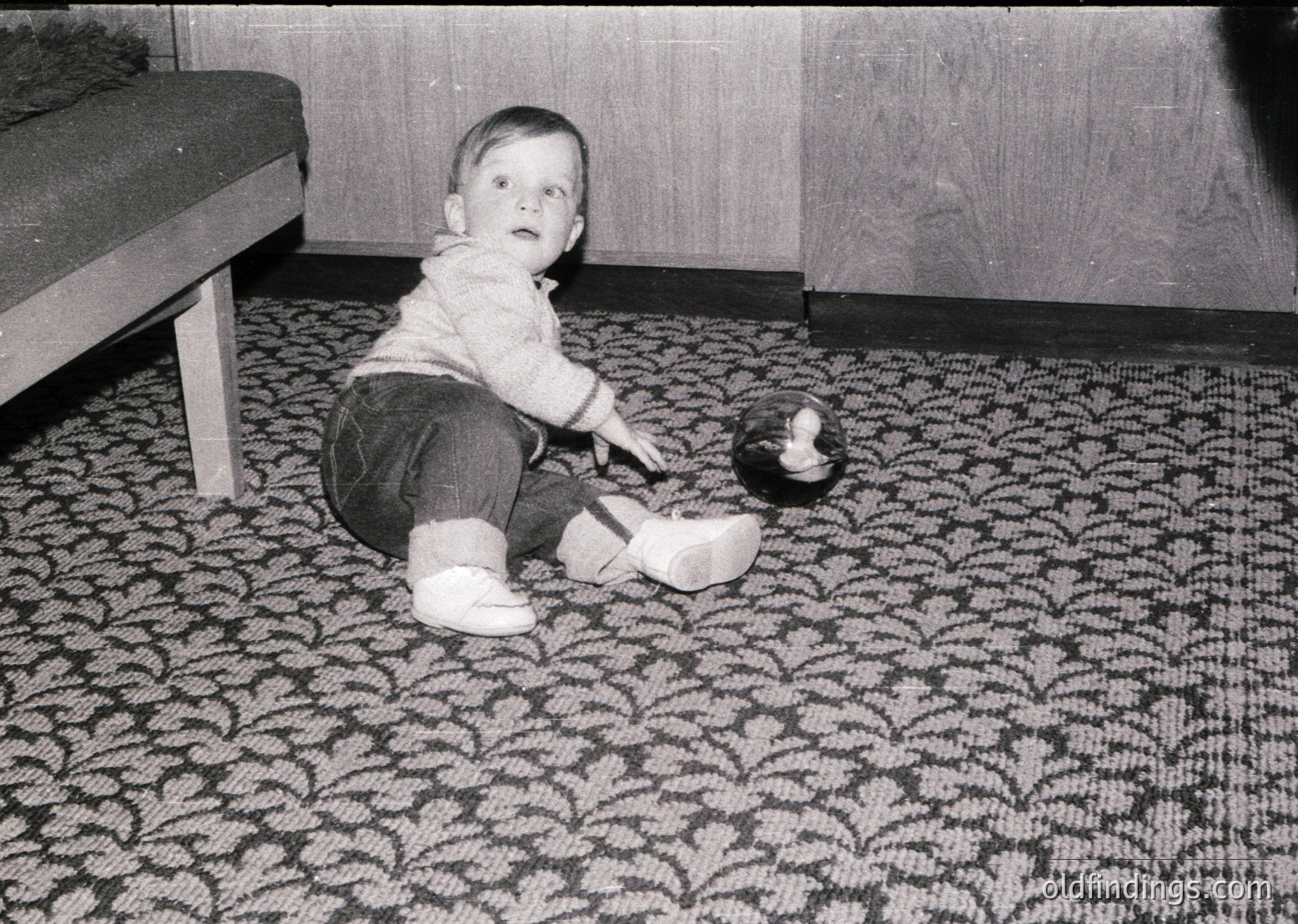 Mid-century indoor scene: toddler in rolled-up trousers and slip-on shoes plays with a small toy duck on a patterned rug, 1950s–1960s. Wooden furniture and simple decor suggest a modest home setting.