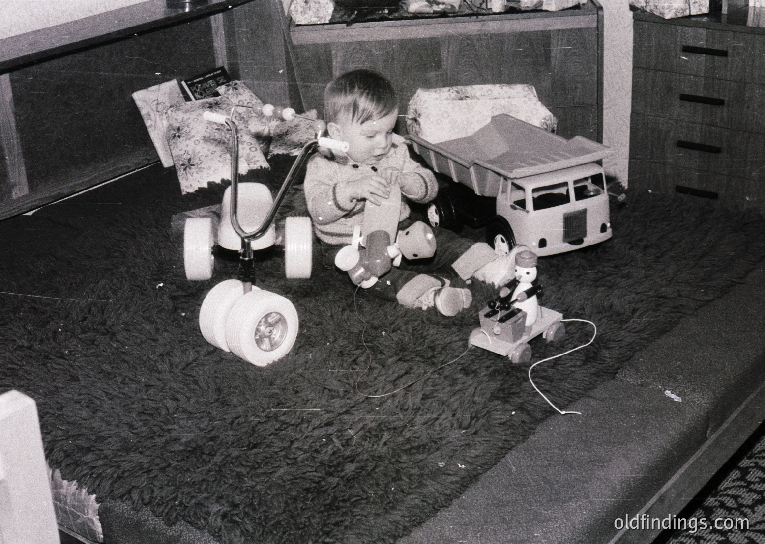 A toddler plays with vintage toys—steam-powered toy train set and toy truck—on a shag-carpeted floor, mid-1960s. Indoor setting features wooden dresser and shelves with knick-knacks.