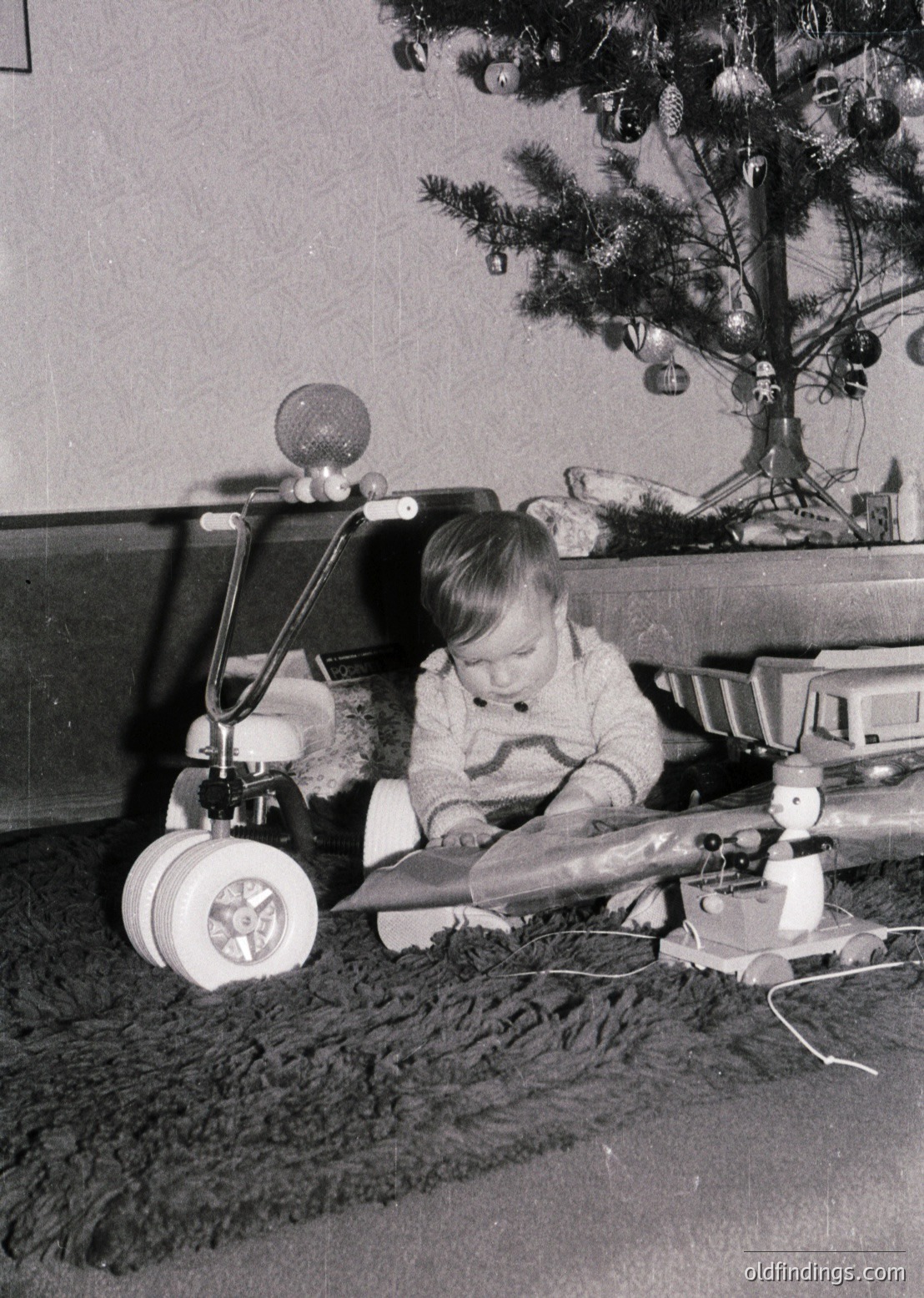Vintage black-and-white photo of a child playing with a toy airplane on a shag rug, beside a decorated Christmas tree with ornaments. Mid-20th century domestic setting, likely 1950s–1960s.