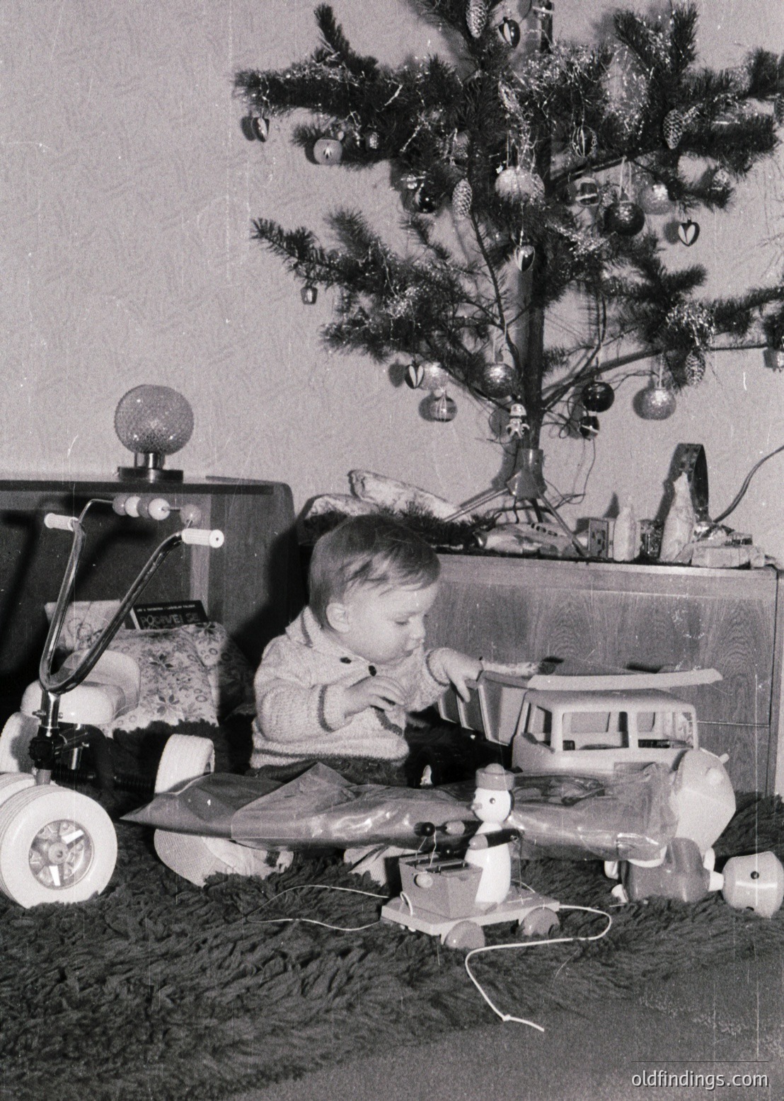 Young child playing with vintage toys—steam train set and toy airplane—on a shag-carpeted floor beside a decorated Christmas tree adorned with baubles and tinsel. Mid-20th century (1950s–1960s) home interior, likely Western.