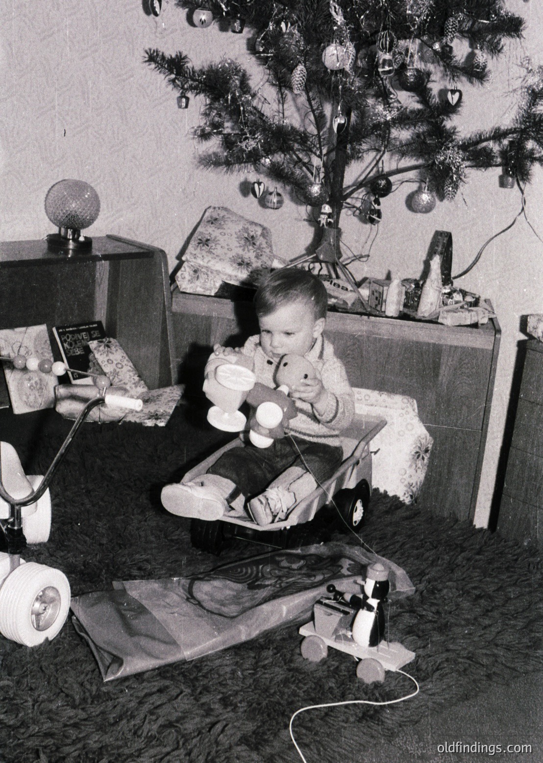 Vintage black-and-white photo of a child in a highchair beside a modestly decorated Christmas tree, holding a toy. Surrounding objects include a radio, wrapped gifts, and a toy truck. Mid-20th century domestic setting.