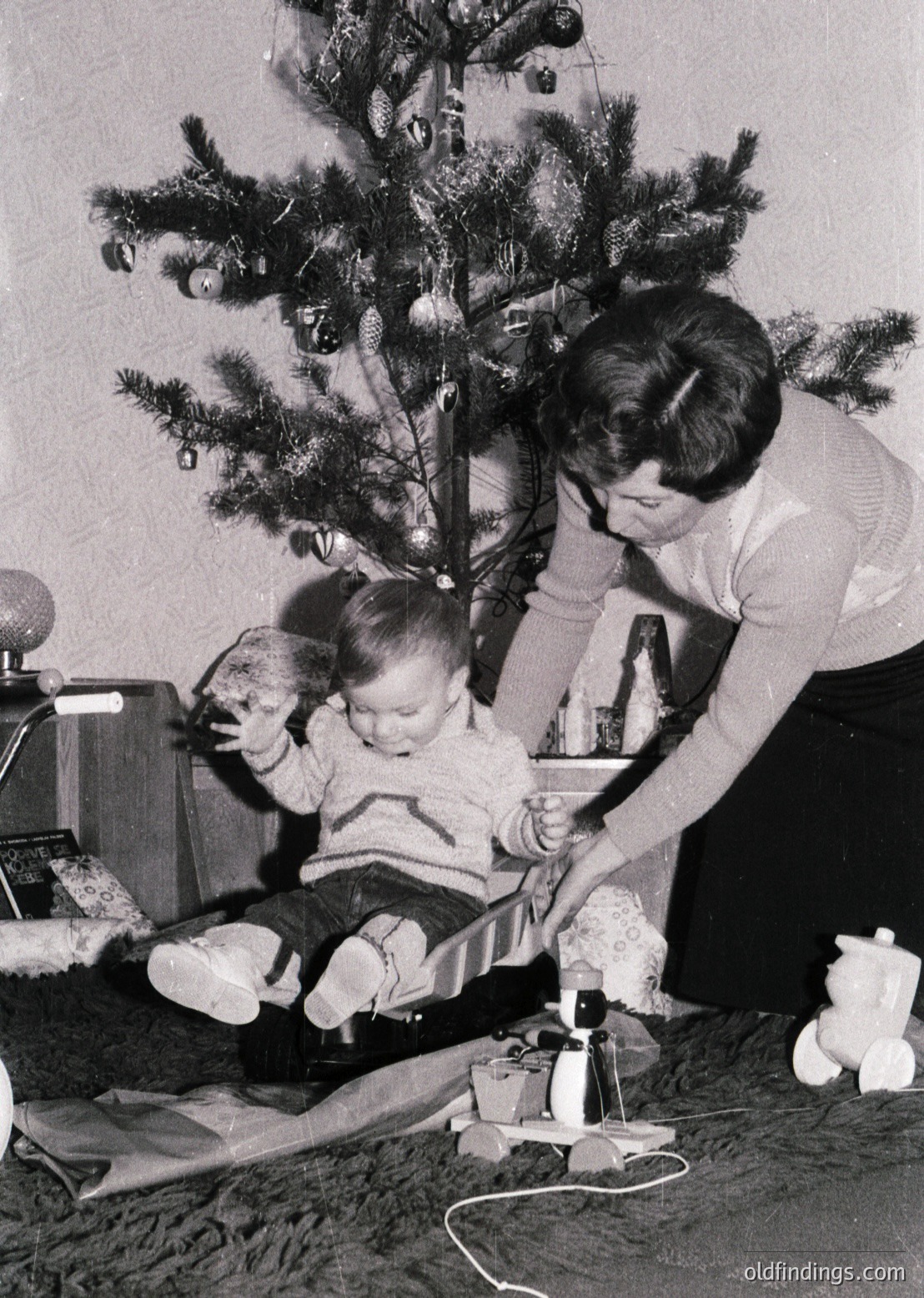 A mid-20th-century (likely 1960s–70s) indoor scene captures a woman decorating a Christmas tree with a child seated on the floor. The tree features tinsel, baubles, and a star topper. The woman holds a candlelit ornament holder, while the child reaches toward a wrapped gift. A vintage radio and a teddy bear add to the nostalgic holiday atmosphere.