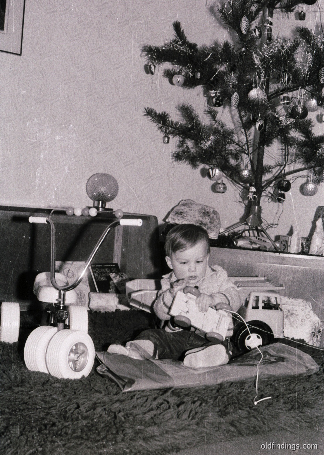 Mid-20th century indoor scene: toddler seated on floor beside vintage toy train set with round wheels and metal track, while a decorated Christmas tree adorns the background.