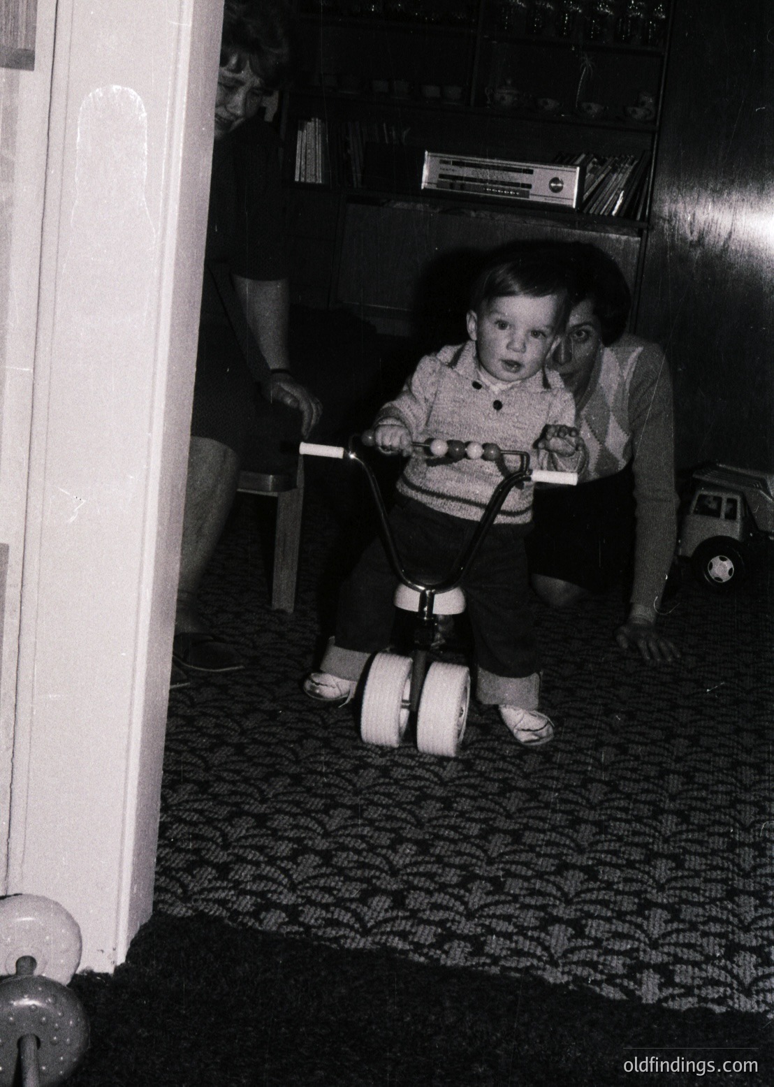 Young child in mid-1970s indoor setting, riding a toy tricycle with training wheels in a dimly lit room. Bookshelves filled with paperbacks and vinyl records in background. Patterned carpet and vintage furniture suggest mid-century domestic life.