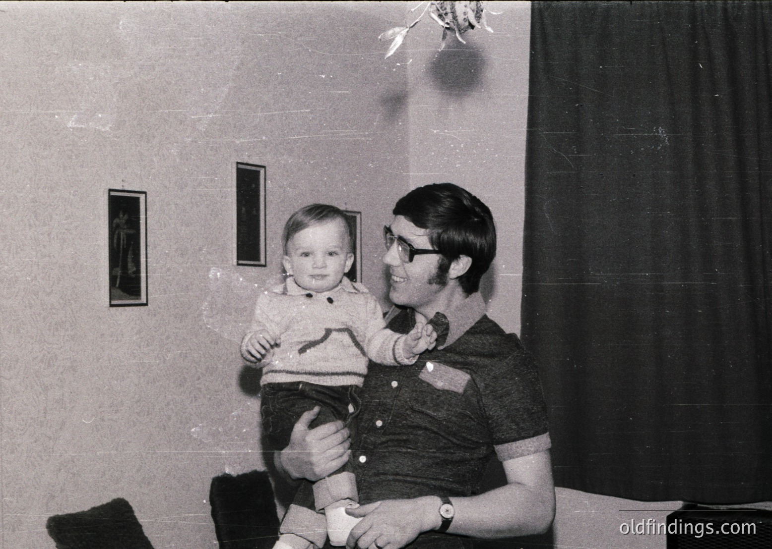 Black-and-white indoor portrait from the 1970s–1980s, featuring a man in a striped polo shirt holding a toddler in a striped sweater. Plain walls with framed photos and a curtained window suggest a modest residential setting. Candid, warm family moment.