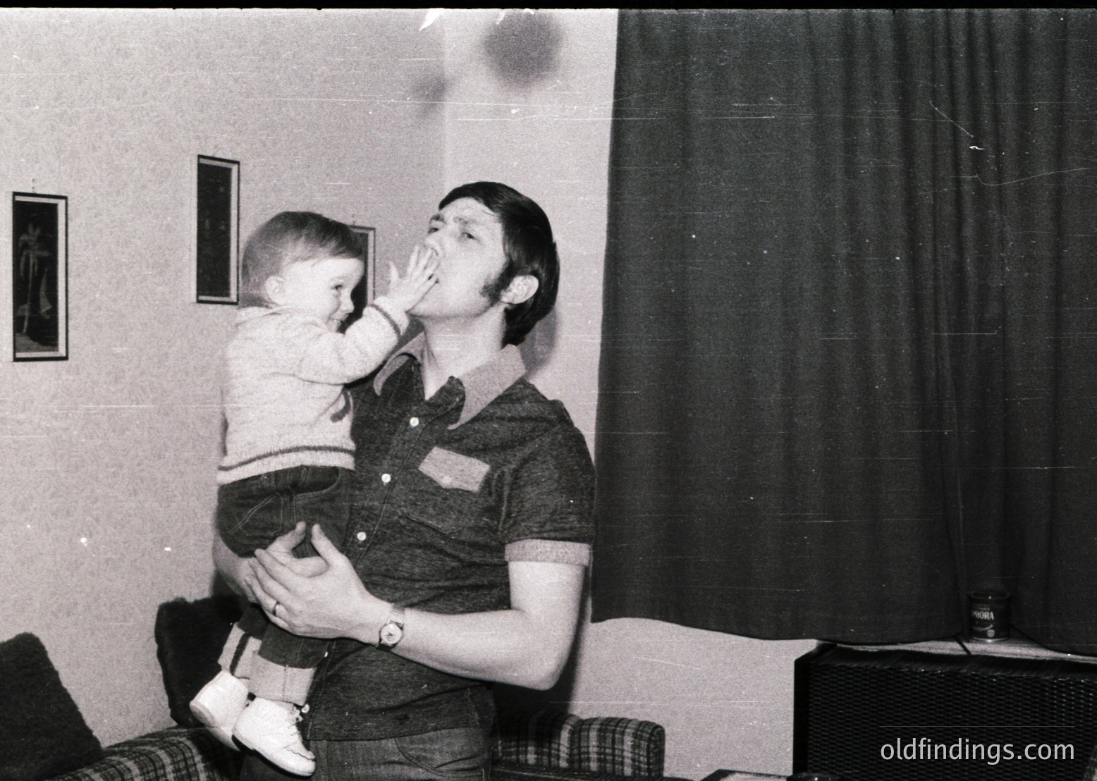 A man in a short-sleeved button-up shirt and jeans playfully kisses a young child indoors, likely mid-1970s. The setting features simple decor: a curtained window, framed art, and a side table with a glass object. Warm, candid family moment.