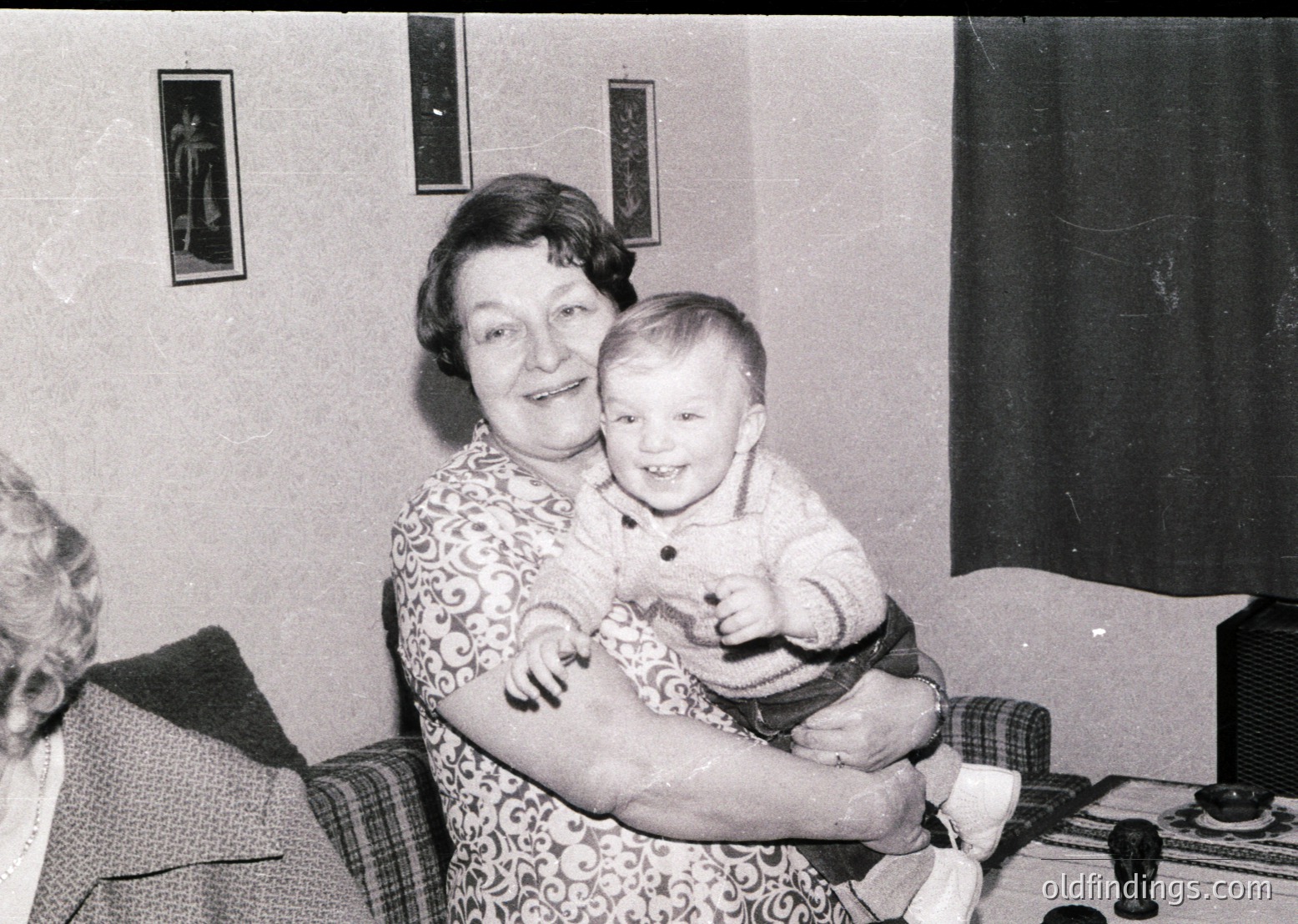 A mid-20th century indoor portrait featuring an elderly woman holding a toddler in a living room setting. The woman wears a floral-patterned dress, while the child is dressed in a light sweater and dark pants. Framed artwork adorns the wall, and a vintage radio or speaker is visible in the background.