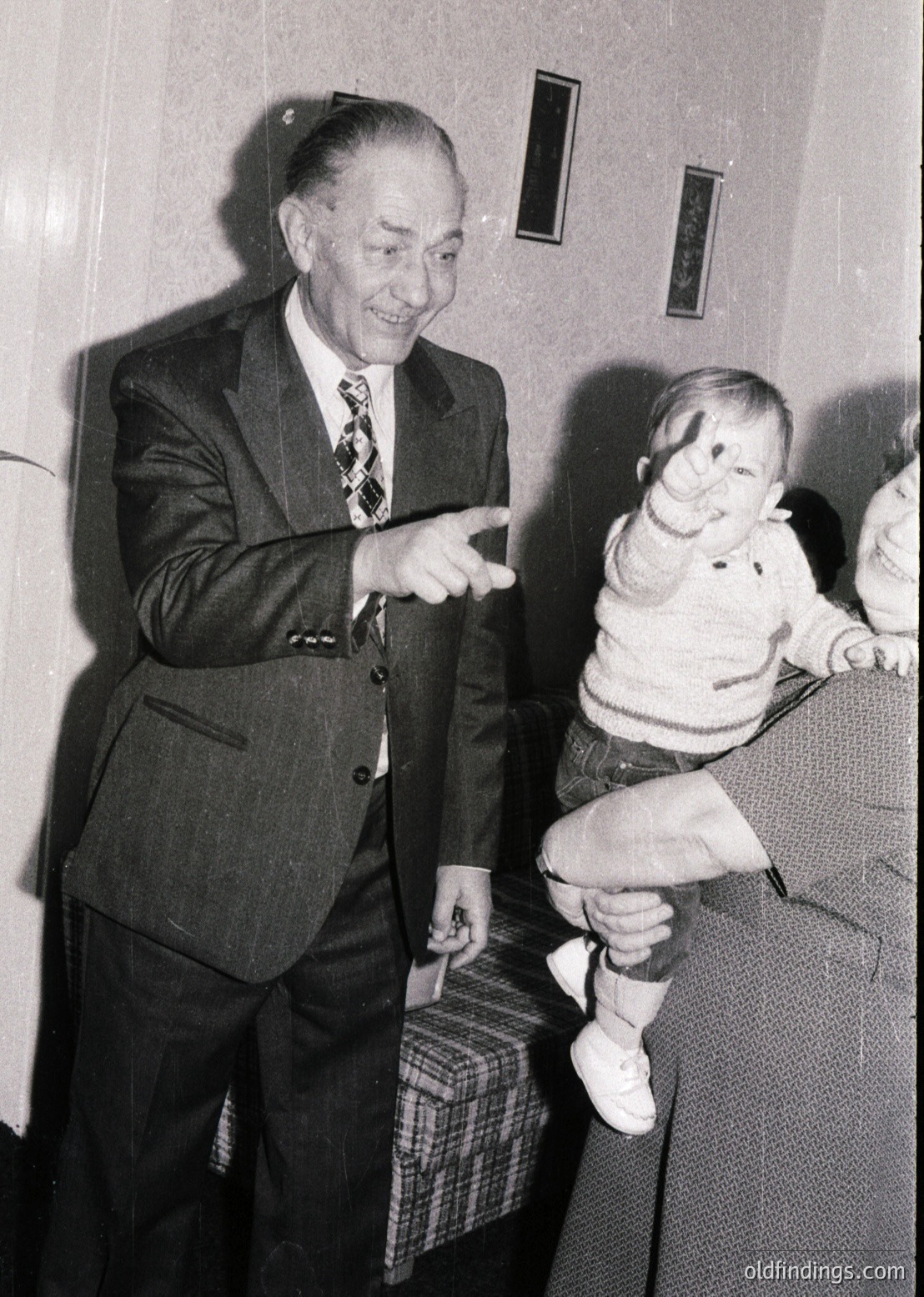 Mid-20th century black-and-white photo of an elderly man in a dark suit and patterned tie playfully lifting a toddler in a sweater and denim overalls. Indoor setting with patterned wallpaper and a checkered blanket on a couch. Candid moment capturing generational bond.