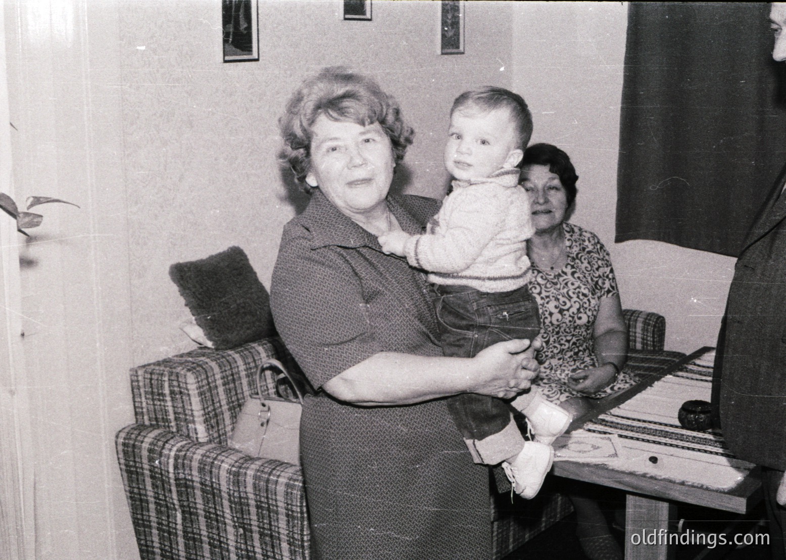 Mid-20th century indoor portrait: woman in 1960s-style dress cradles a baby, flanked by another seated woman in patterned blouse. Checkered sofa and vintage piano suggest a mid-century home setting. Warm familial moment captures generational bonds.