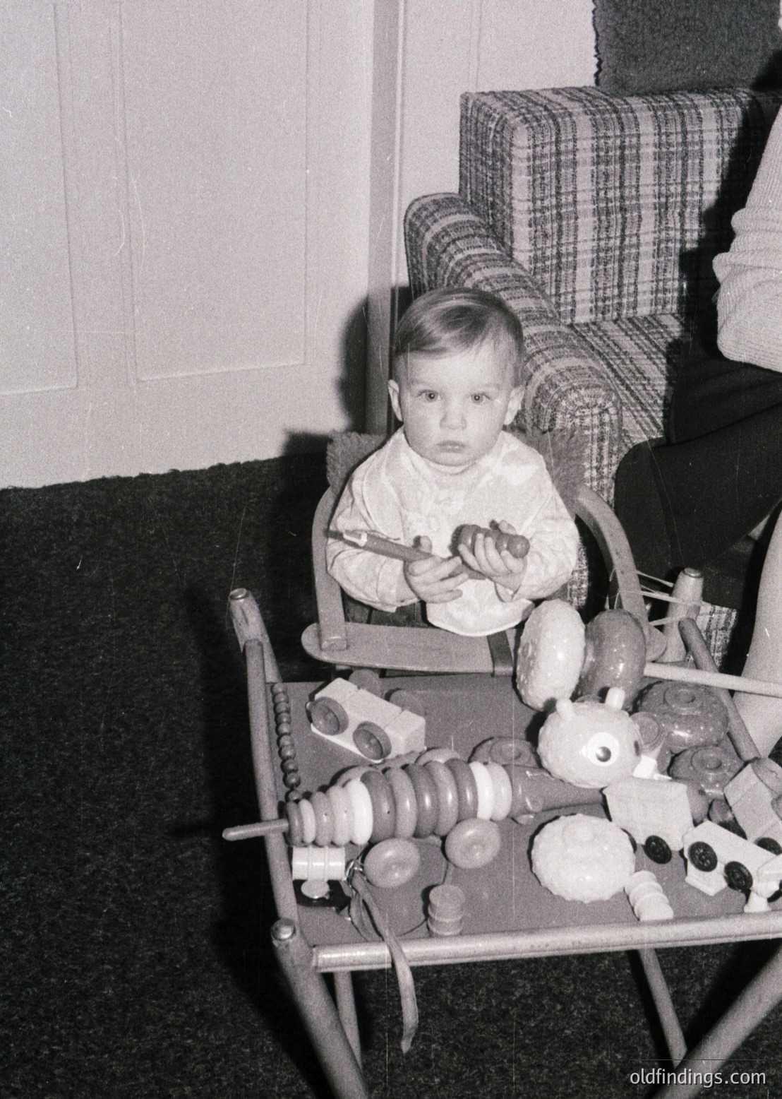 Vintage black-and-white photo of a toddler seated in a highchair, holding a spoon toward a tray filled with classic wooden toys: blocks, animal figurines, and round knobs. Plaid upholstered chair in background suggests mid-20th century domestic setting.