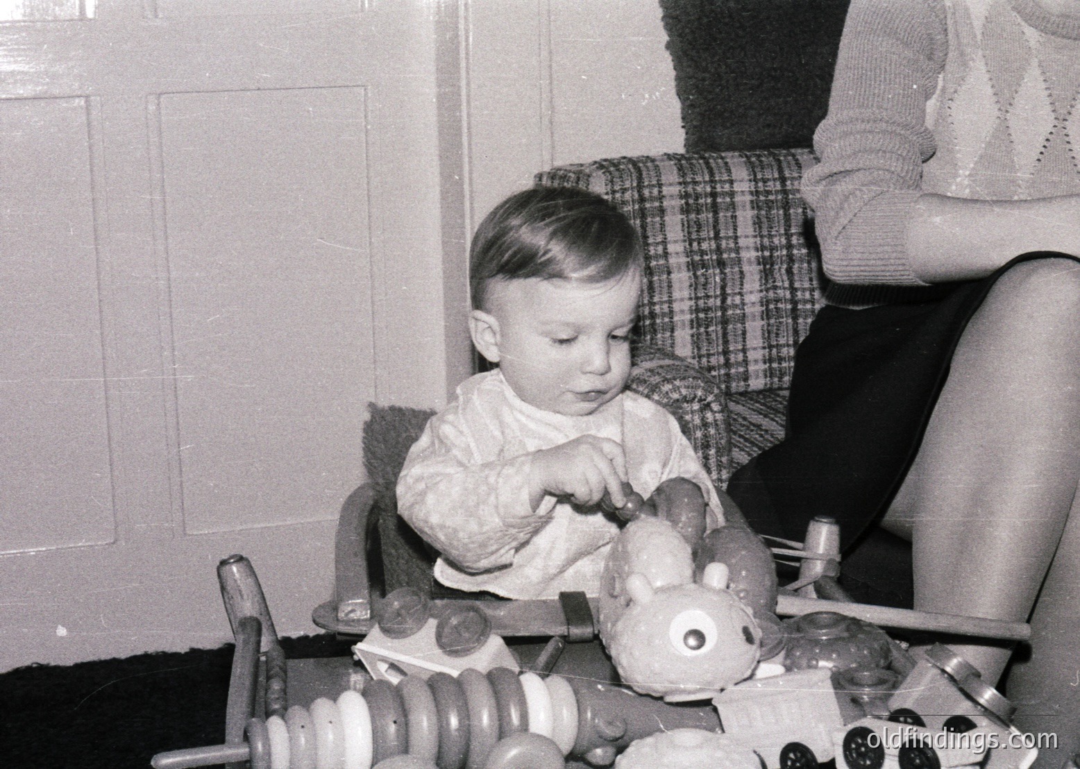 Toddler in 1960s-style sweater plays with vintage toy train set on wooden table. Adult’s plaid skirt and knit sweater visible in background. Indoor domestic scene evokes mid-century childhood nostalgia.