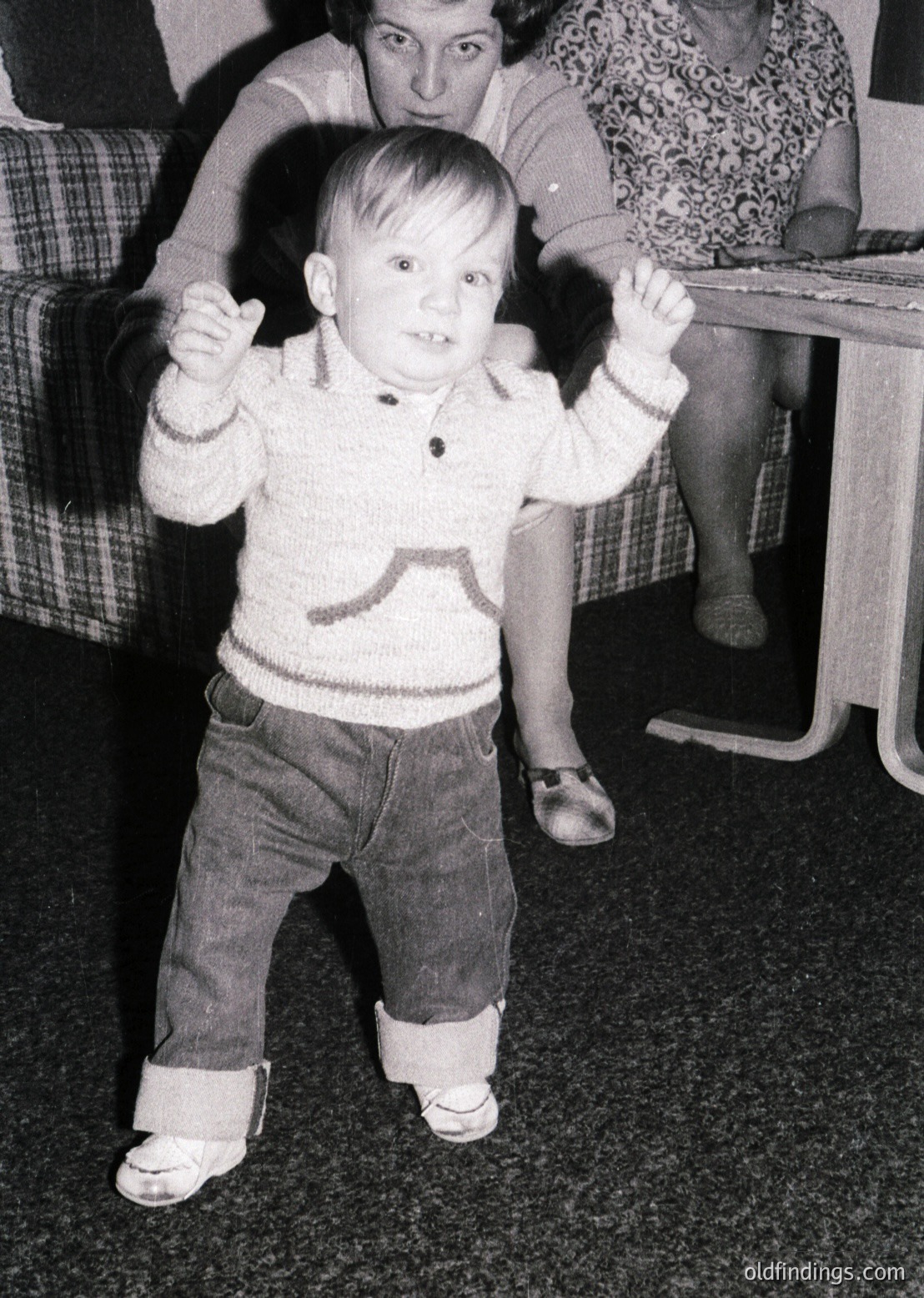 Young child in 1960s-style sweater and knee-high socks, mid-dance pose with arms raised. Indoor setting with patterned sofa and wooden furniture. Candid moment capturing playful energy.