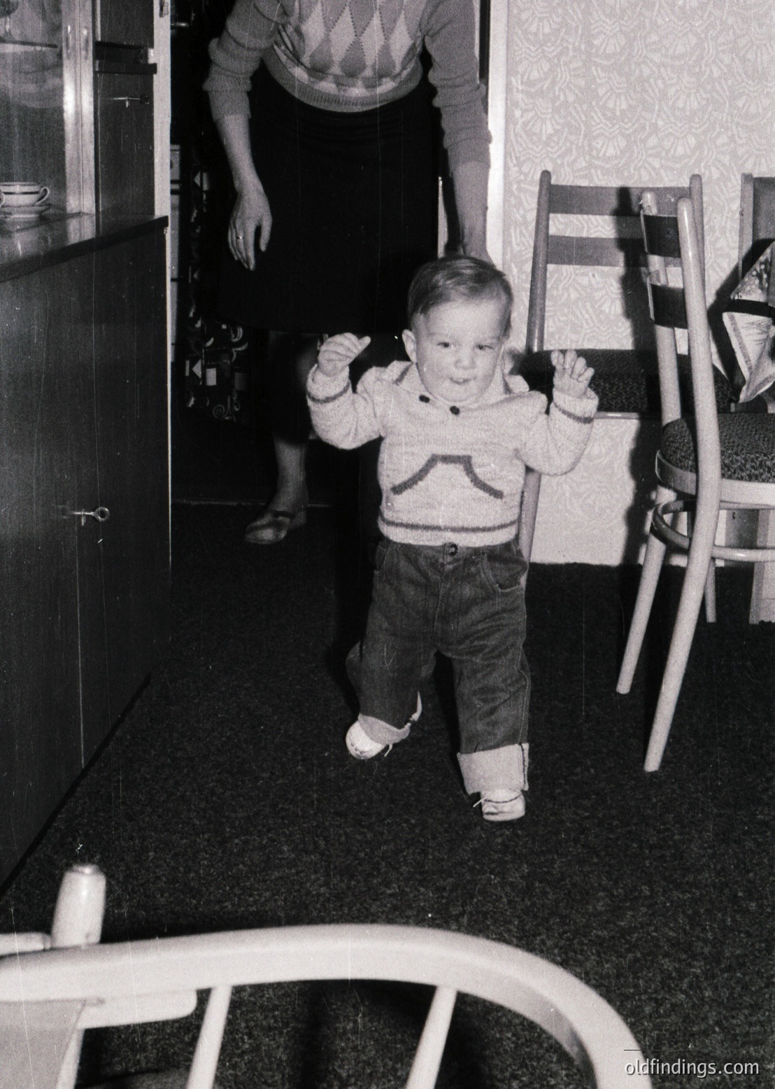 Toddler in mid-stride on dark carpeted floor, wearing 1960s-style sweater and denim pants with white shoes. Adult partially visible in doorway, holding kitchen cabinet door. White highchair and patterned wallpaper in background. Candid, mid-century domestic scene.