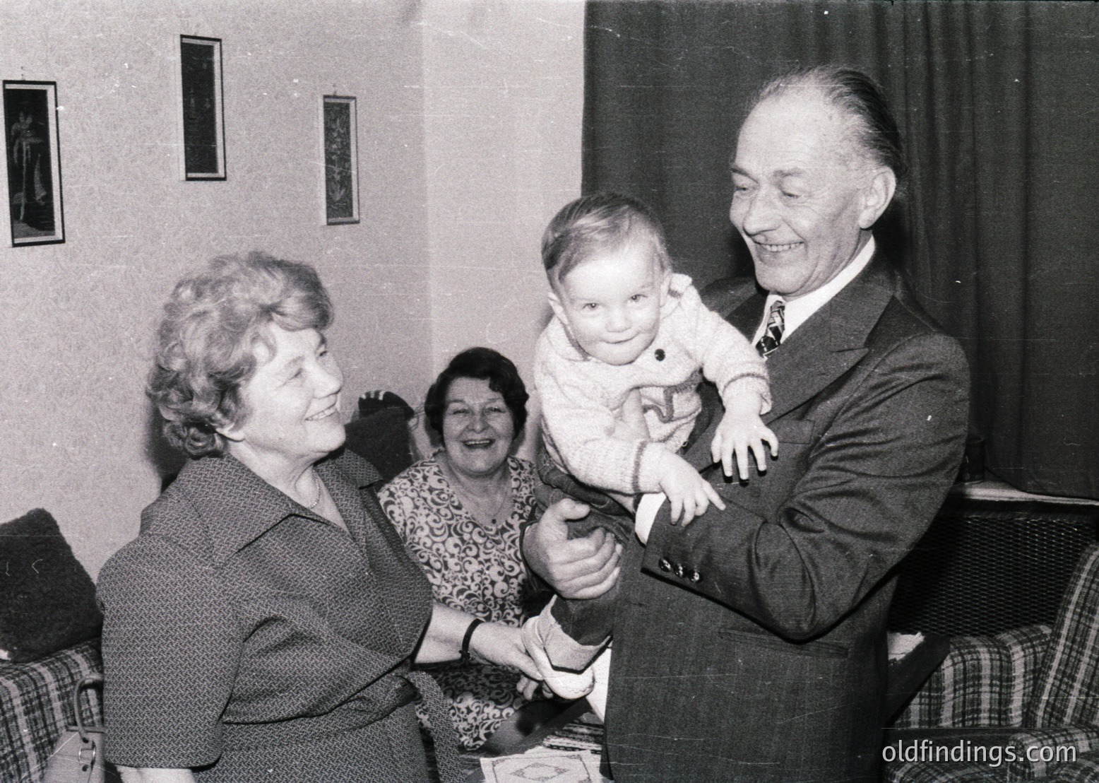Intergenerational gathering in a mid-20th-century living room, likely 1950s–1960s. Elderly man in suit cradles a baby, while a woman in floral dress and older woman in a dark dress smile. Plain curtains, framed art, and plaid sofa suggest modest domestic setting.